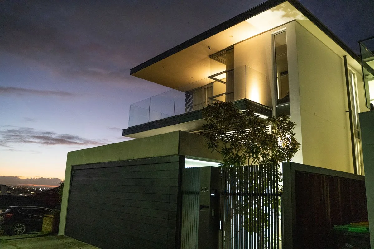 Modern multi-story house at dusk with balcony and large windows, illuminated exterior lighting, and a mesh gate in the front yard, with a car parked outside.