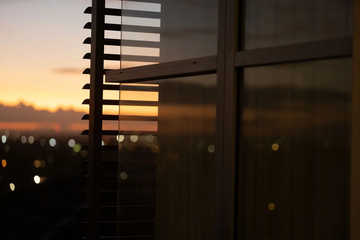 Sunset view through a window with blinds and glass panels, city lights visible in the distance.