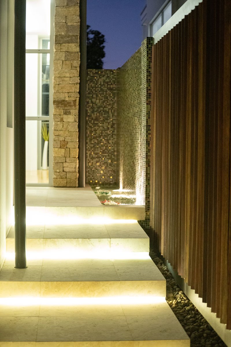 Night view of an outdoor modern patio area with illuminated steps, a stone wall, a mosaic tile wall, and a wooden fence.
