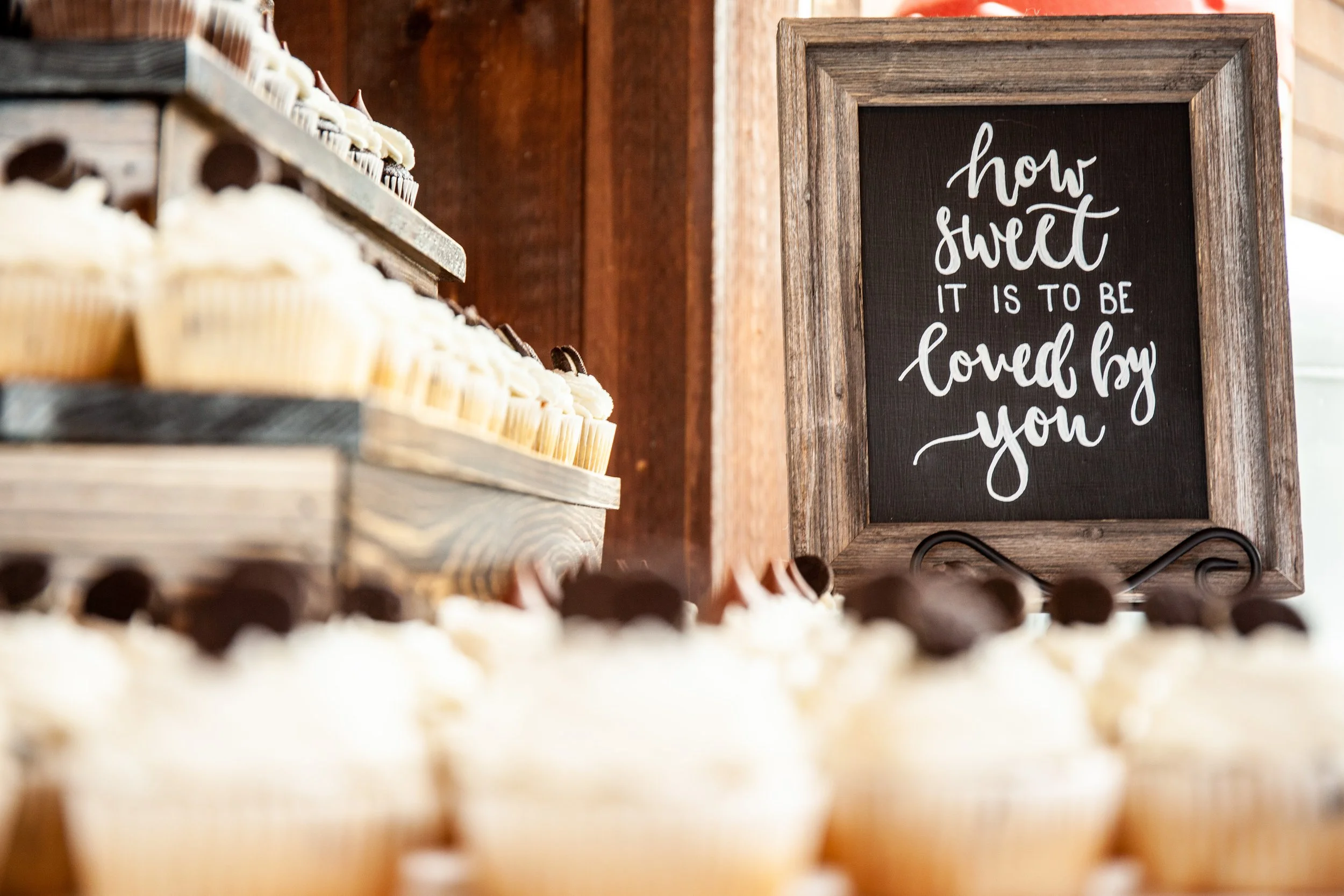 A display of cupcakes with vanilla and chocolate frosting on wooden shelves, with a framed sign in the background that says, "how sweet it is to be loved by you."