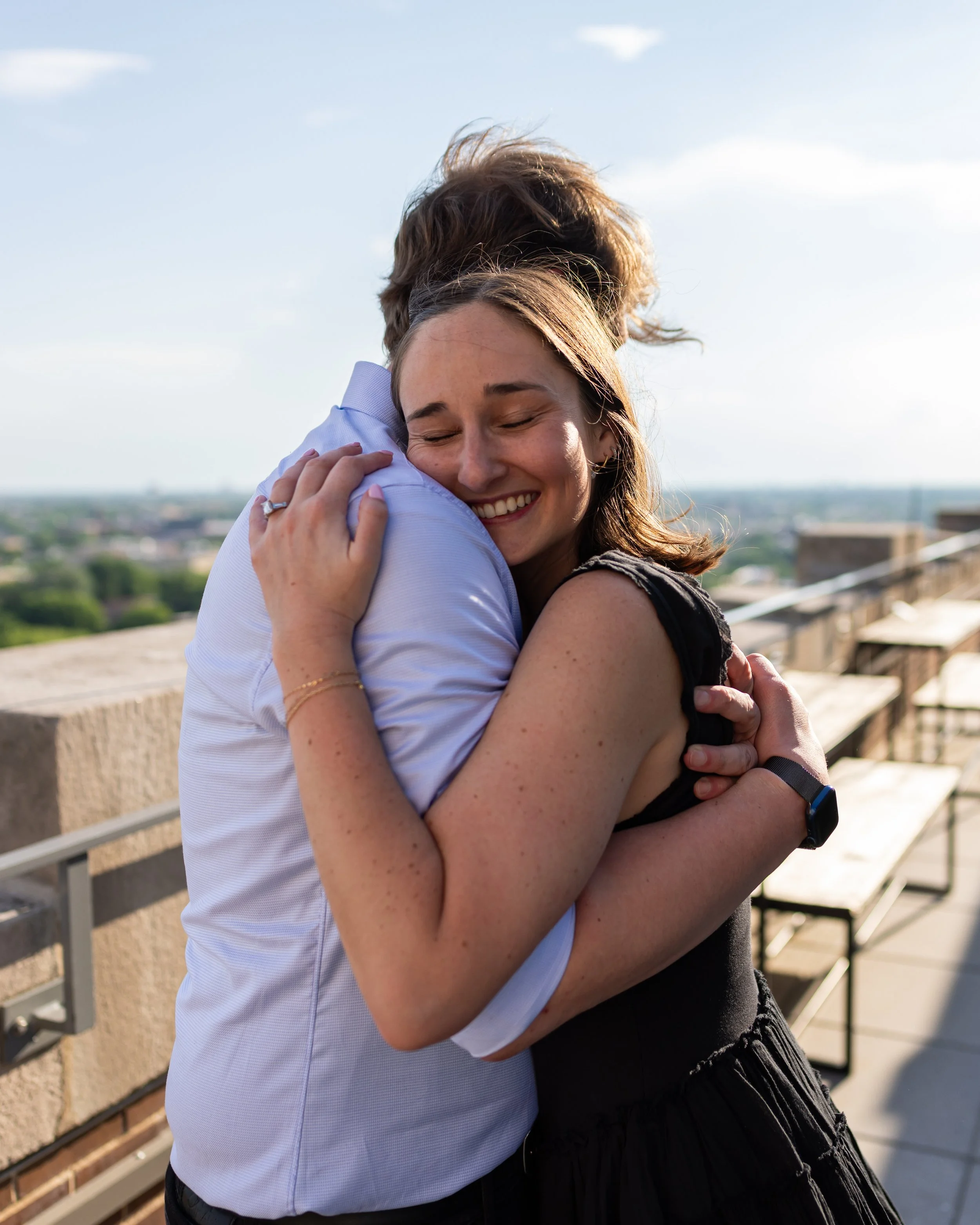 Two women embracing and smiling outdoors on a rooftop balcony, with a cityscape and blue sky in the background.