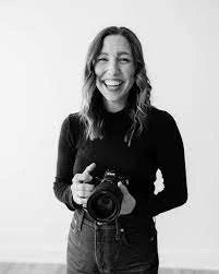 A woman smiling and holding a camera, wearing a black top and jeans in a black and white photo.