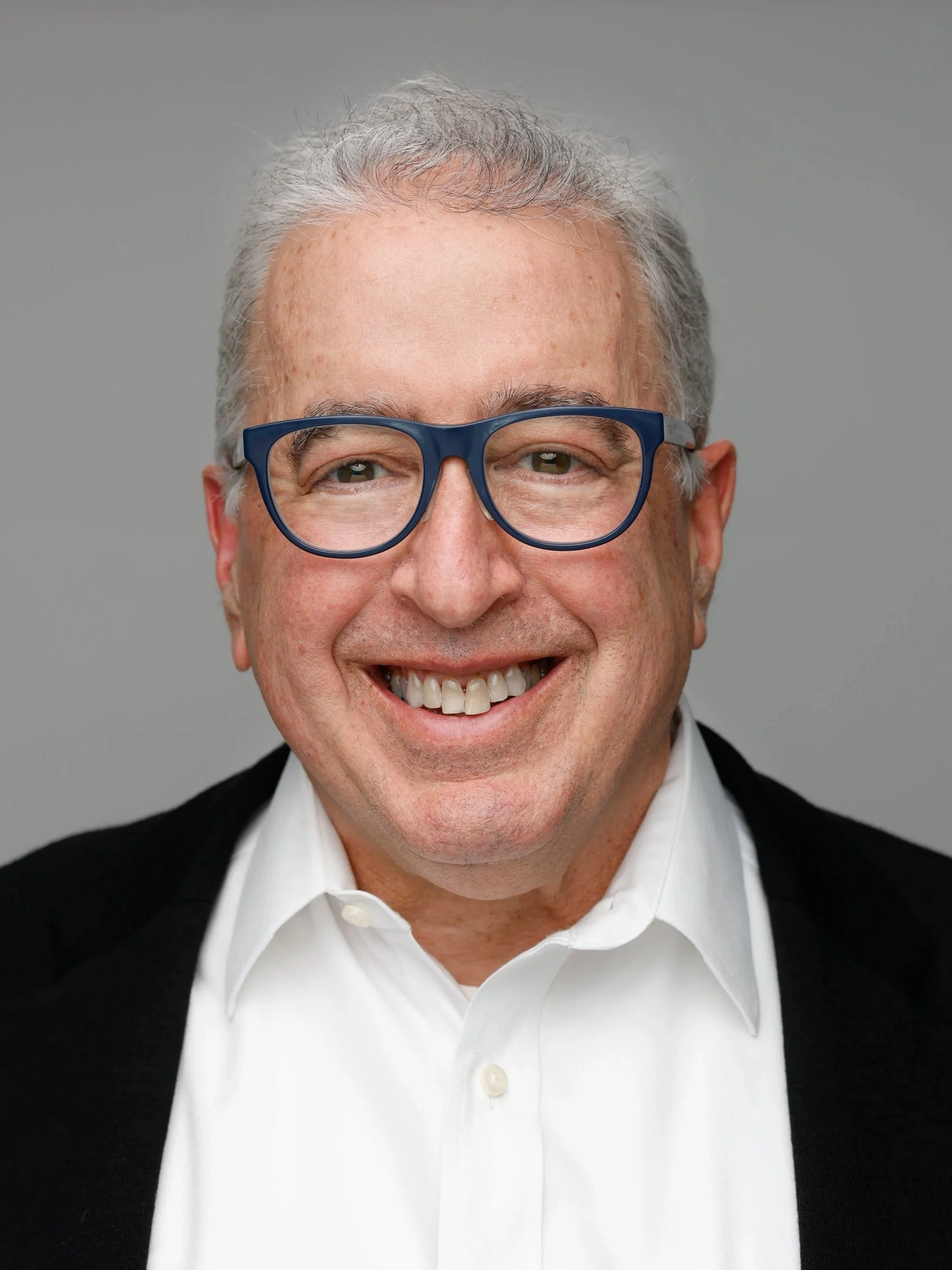 Portrait of a smiling middle-aged man with gray hair, wearing glasses, a white shirt, and a dark blazer against a gray background.