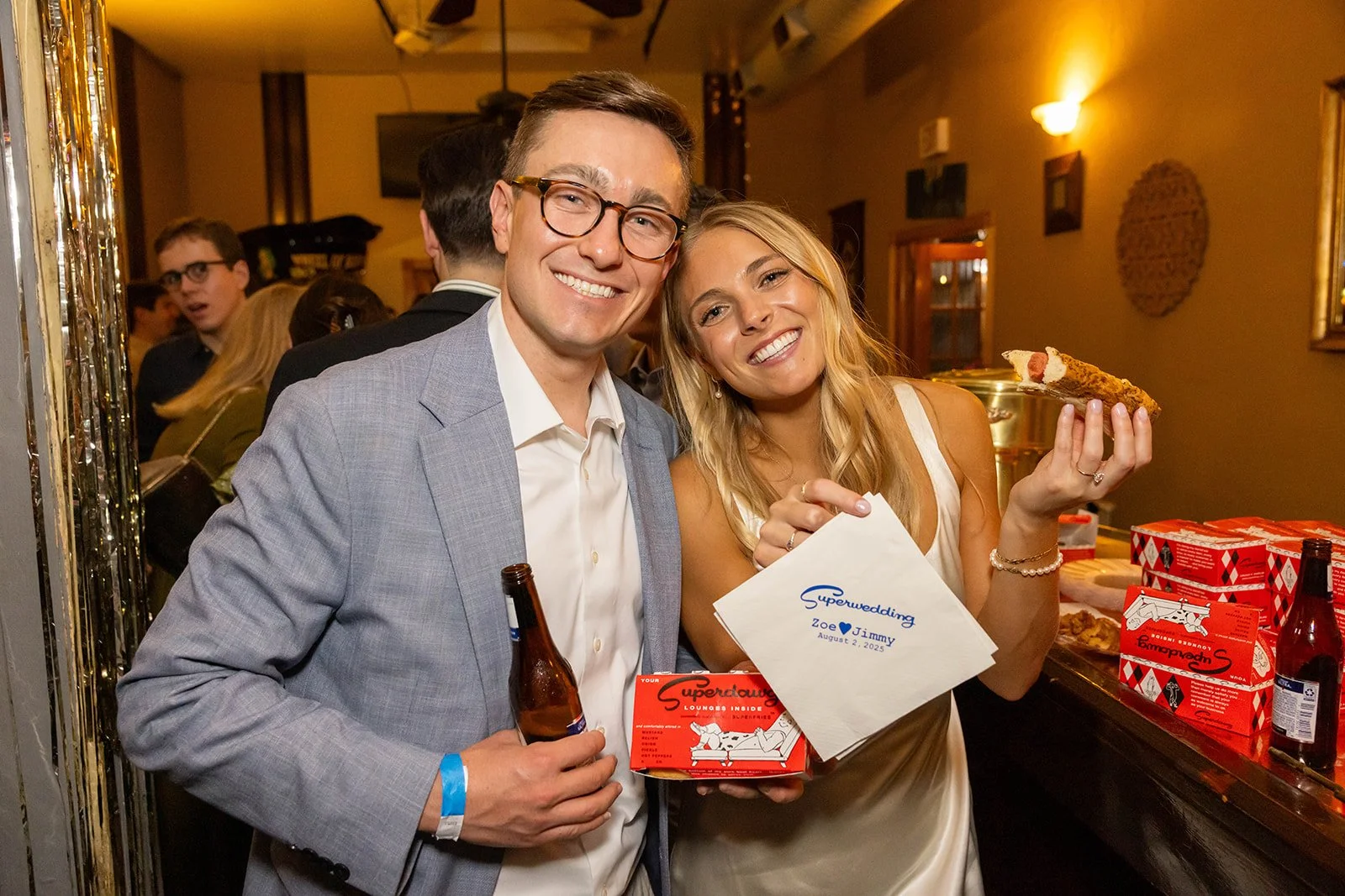 A smiling man in glasses wearing a gray blazer and a woman with blonde hair in a white dress holding pizza and a wedding card at a wedding reception.