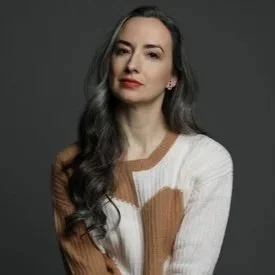 A woman with long wavy dark hair poses against a dark gray background, wearing a cream-colored sweater.