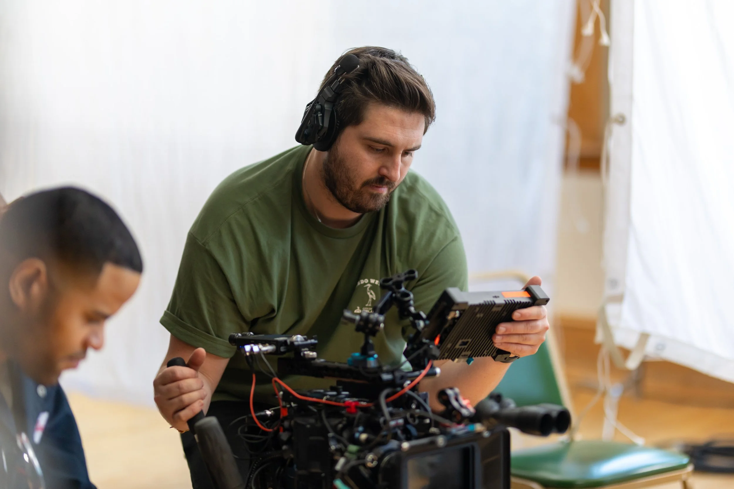A man in a green shirt with headphones operating a film or video camera with visible wiring, while another person is partially visible in the foreground in an indoor setting.