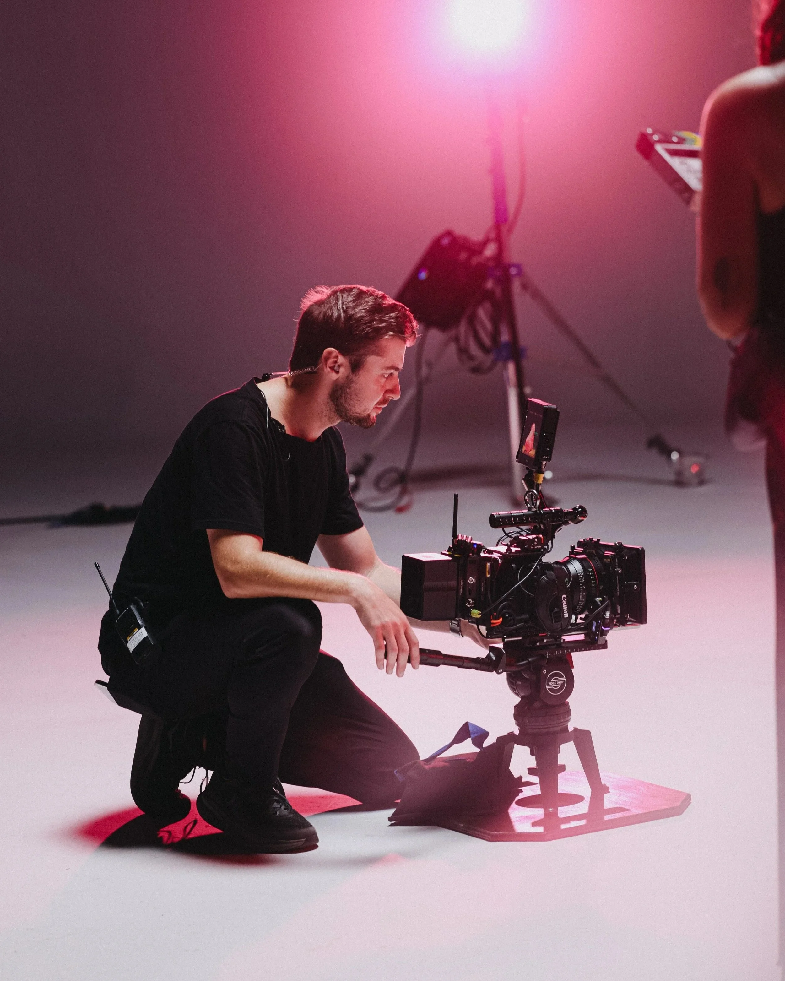 A man kneeling on the floor operating a professional camera on a tripod, with pink lighting and equipment in the background.
