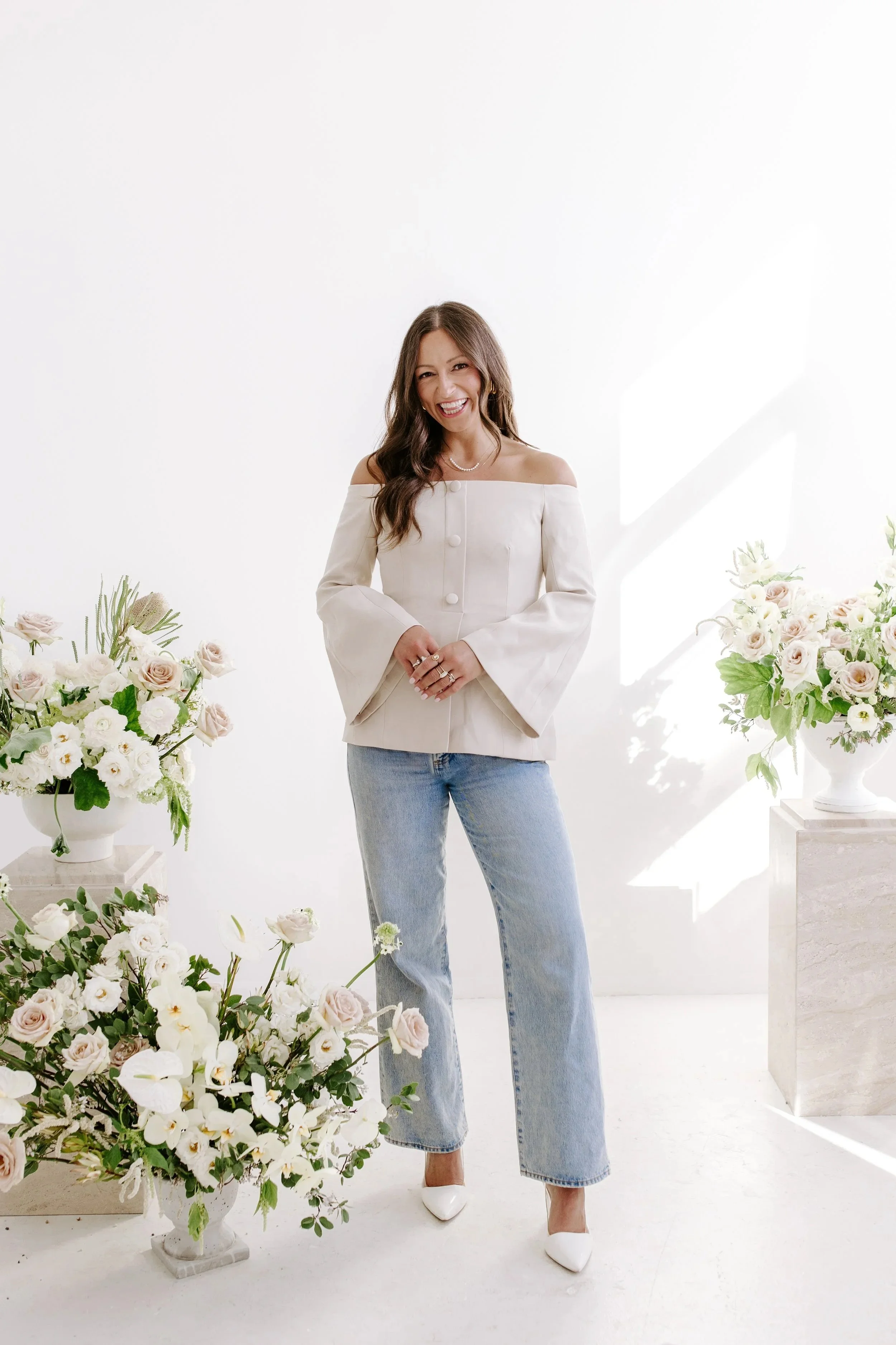 A woman smiling and standing in a bright, white studio surrounded by white and light pink flower arrangements.