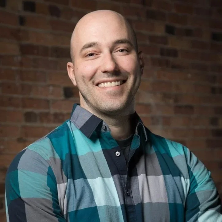 A smiling bald man with light skin, wearing a colorful checkered shirt, stands in front of a brick wall.