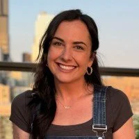 A woman with dark hair smiling, wearing a black top and denim overalls, standing outdoors with a city skyline in the background.