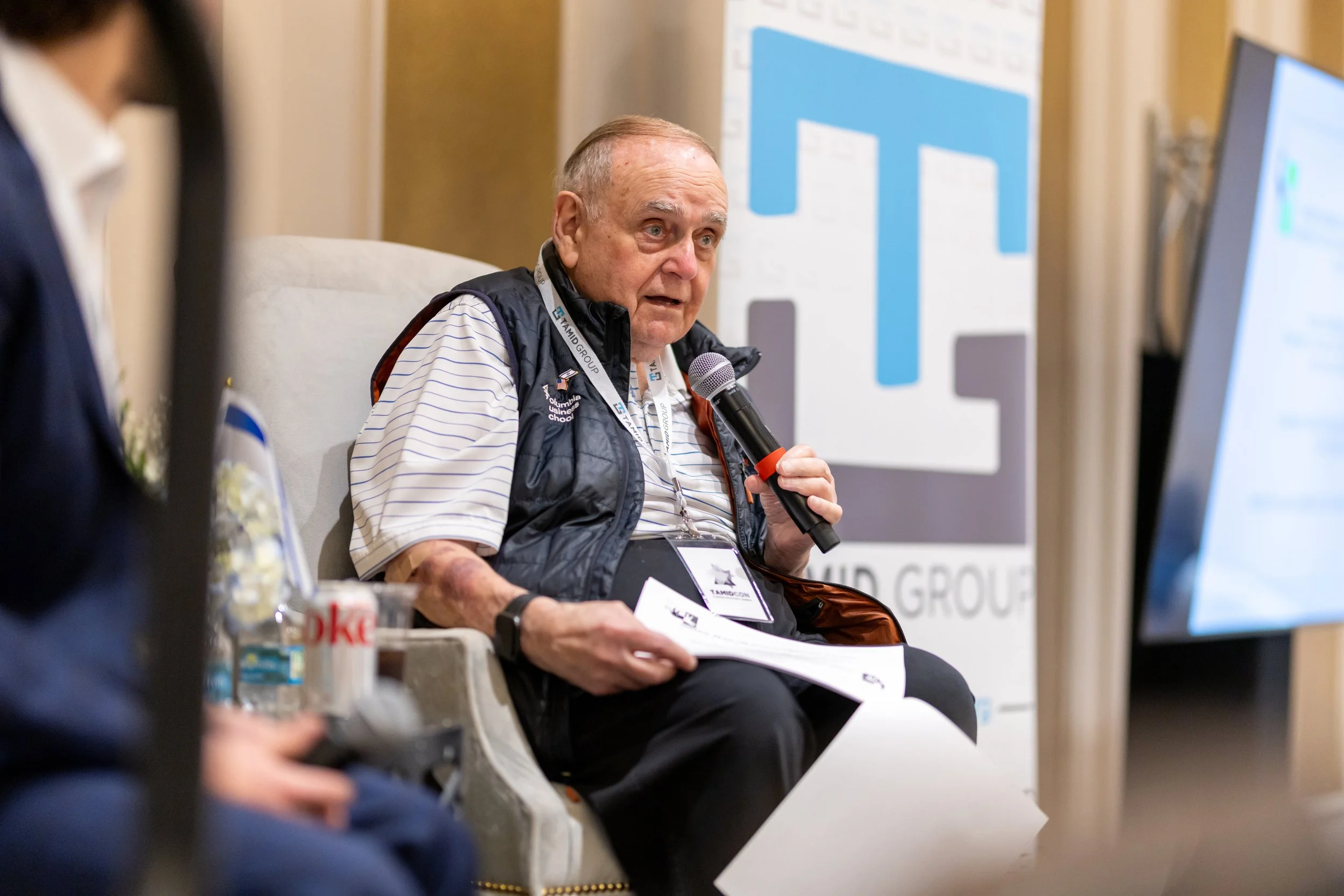 An elderly man is sitting on a bench, holding a microphone, speaking at an event. He is wearing a striped shirt, a black vest, and has a badge around his neck. There is a large monitor screen to his right and a promotional banner in the background.