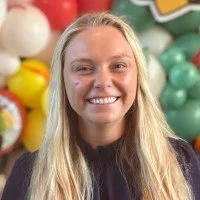 A smiling woman with long blonde hair in front of colorful balloons.