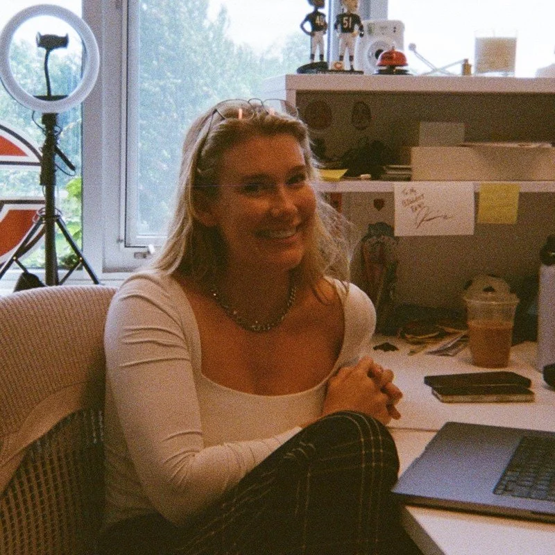 A woman sitting at a desk in a room with various objects and equipment, smiling at the camera.