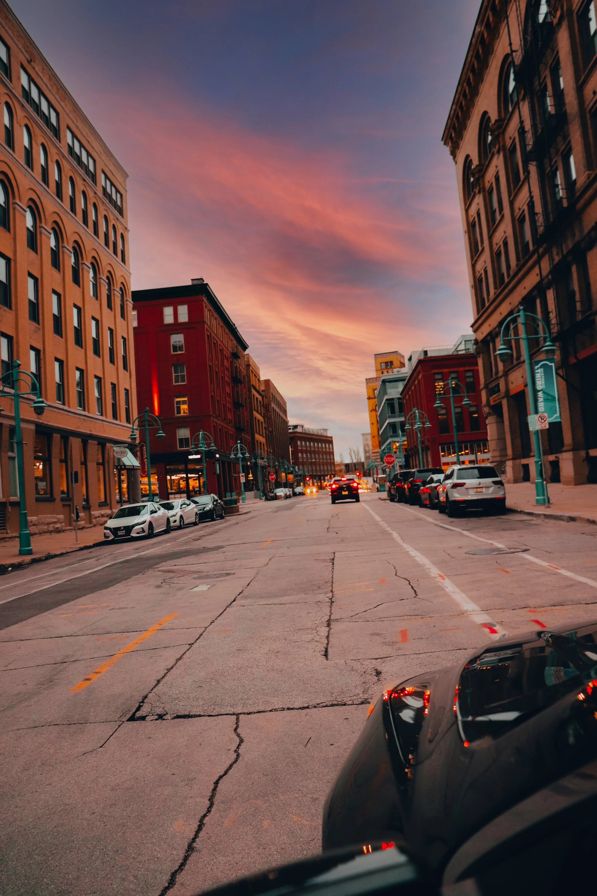 A city street at sunset with colorful sky, tall buildings on both sides, parked cars along the curb, and a few cars driving down the road.