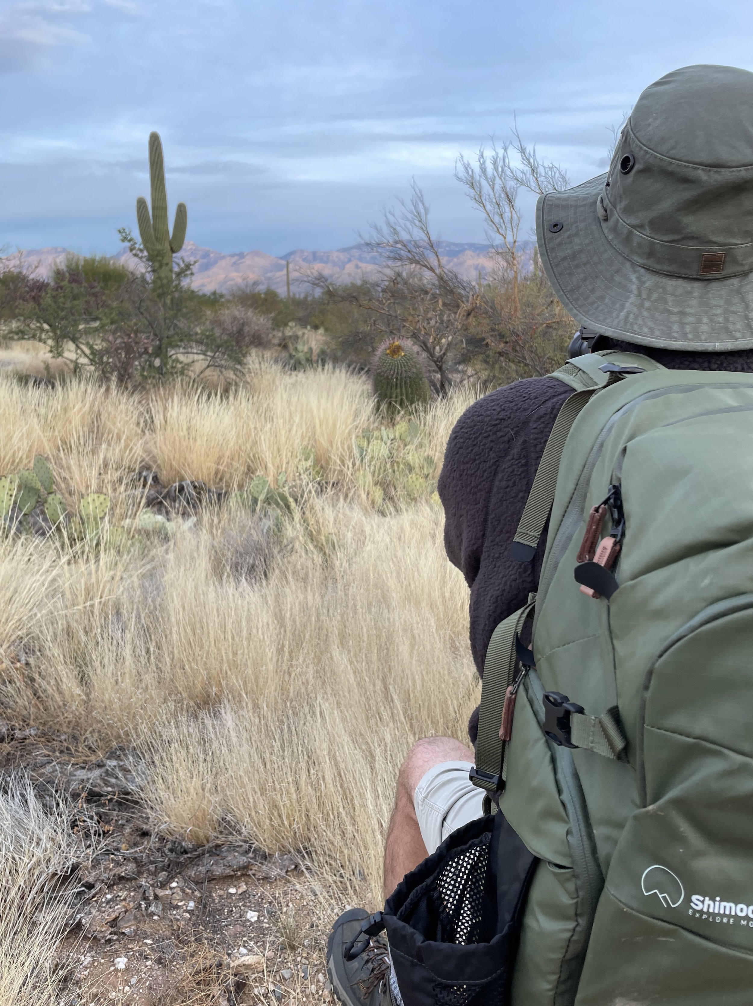 Jeffrey Tadlock wearing a hat and backpack sitting on a trail in a desert landscape, with cacti and mountains in the distance, under a cloudy sky.