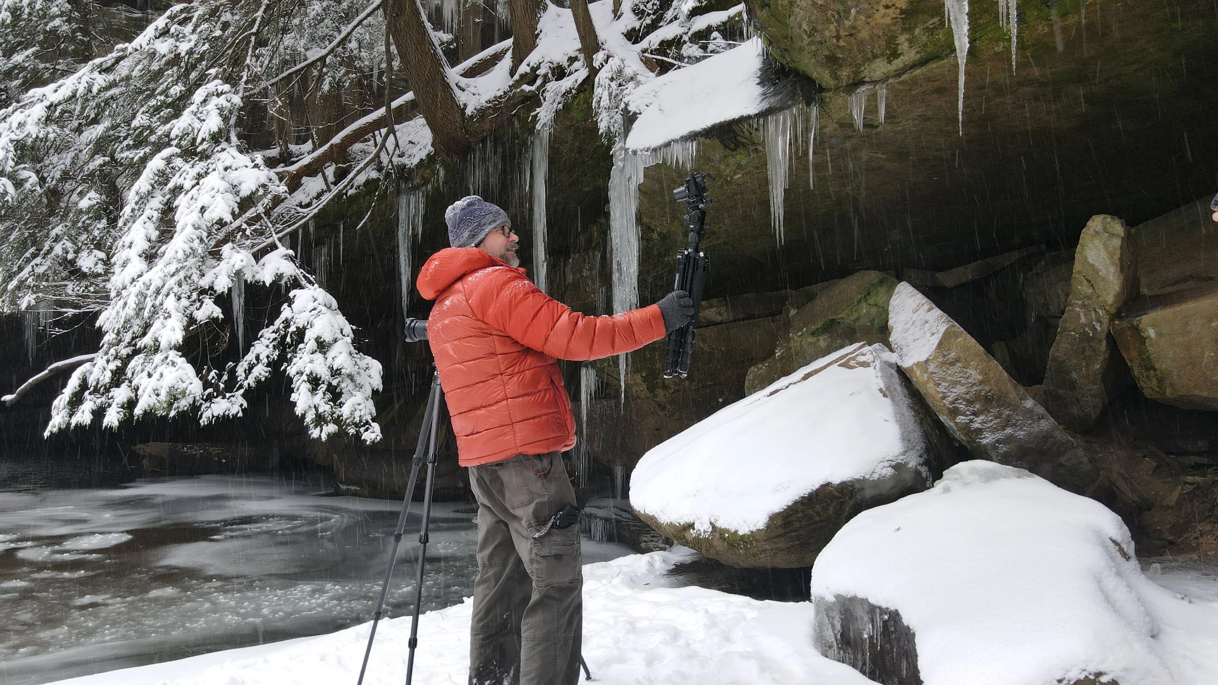 Jeffrey Tadlock in a red jacket and gray beanie taking photos of icy rocks and snow-covered trees in a winter landscape.