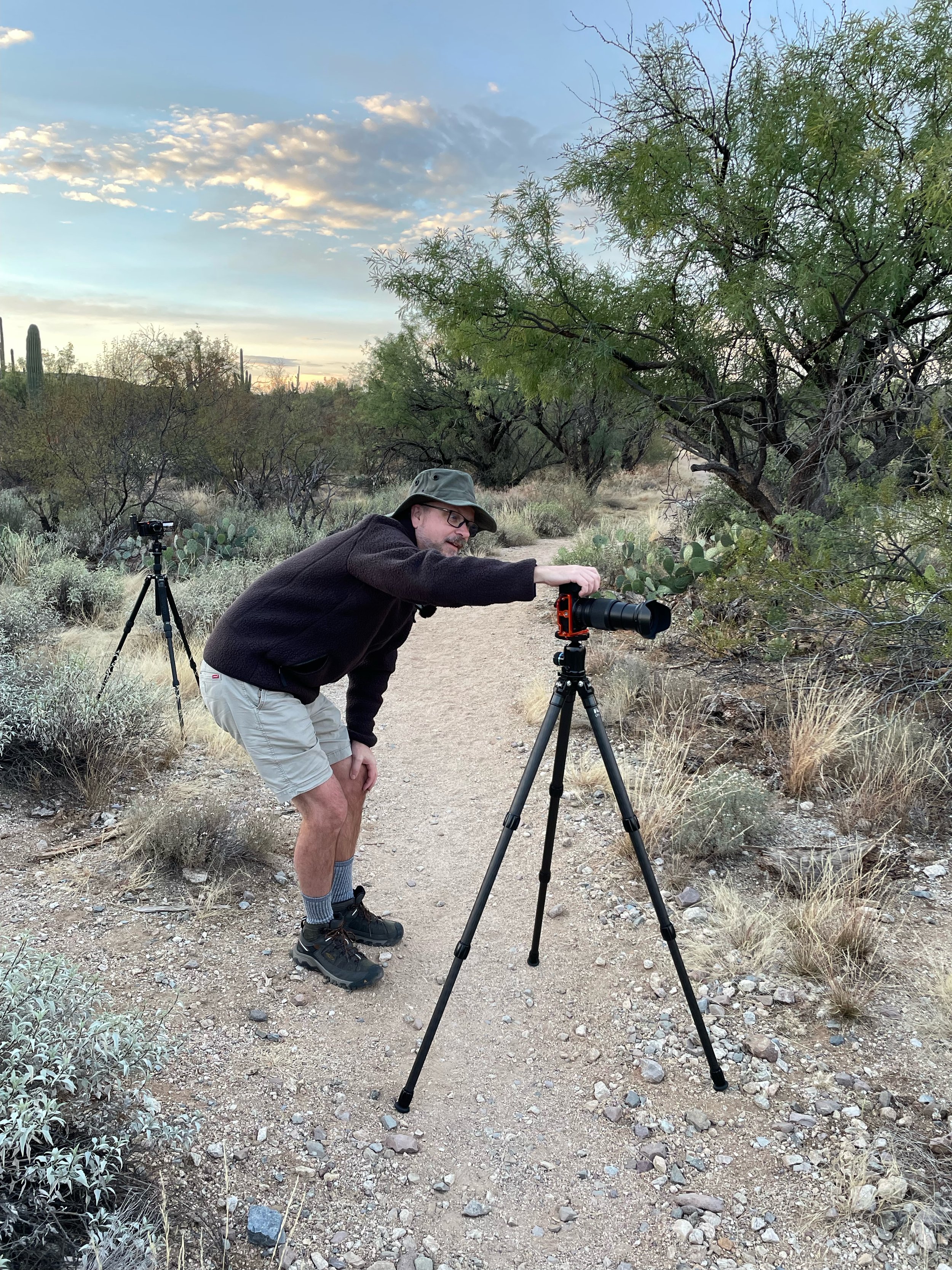 Jeffrey Tadlock, landscape photographer, in outdoor clothing, including a hat, glasses, a black sweater, and shorts, is using a camera on a tripod to take photos in a desert landscape with cacti and bushes, during sunrise or sunset.