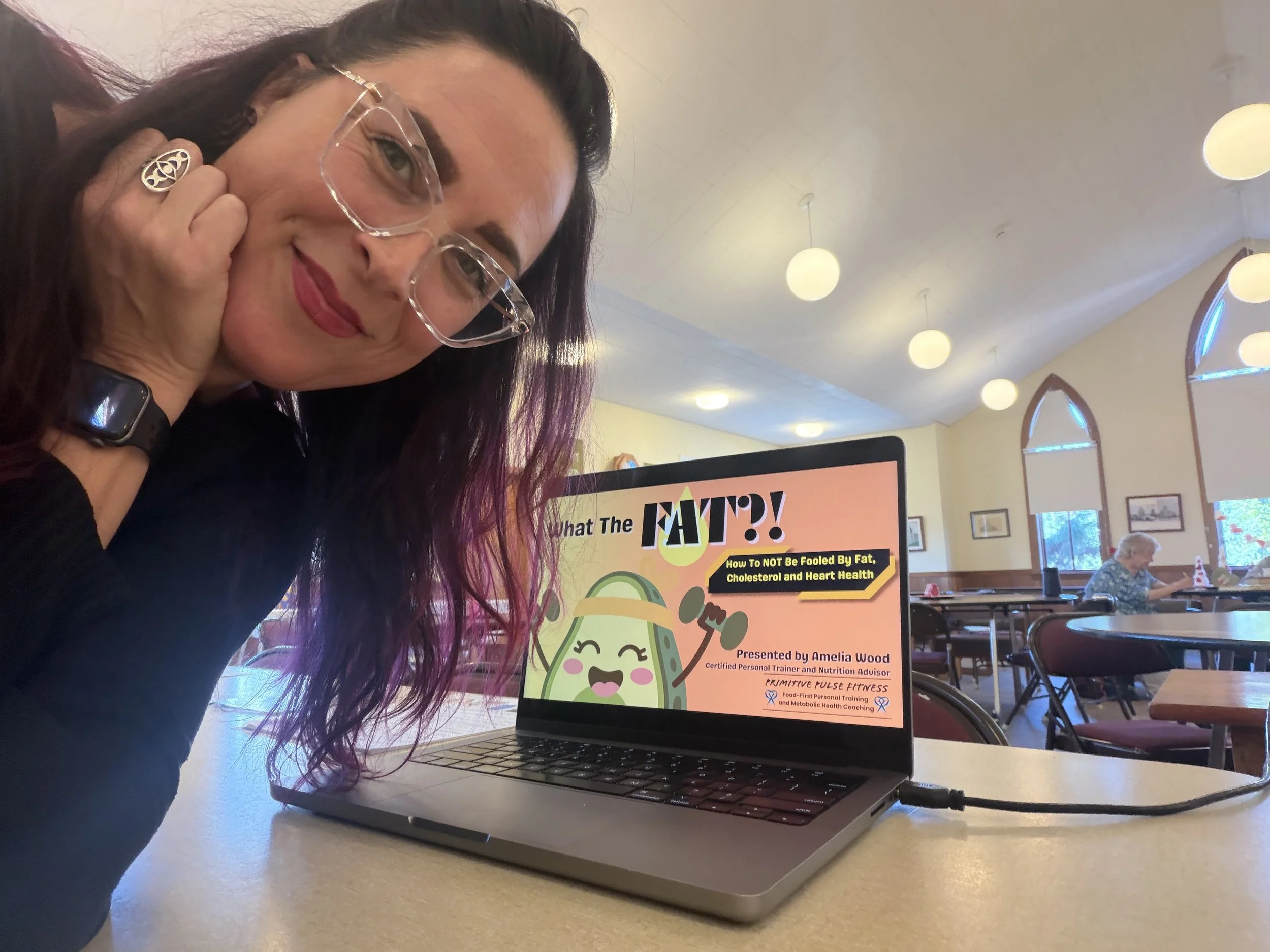 A woman with glasses and purple hair posing next to a laptop in a dining hall. The laptop screen displays a presentation titled "What The FATTY?!" about how to avoid being fooled by fat, cholesterol, and heart health. Other diners and large windows are visible in the background.