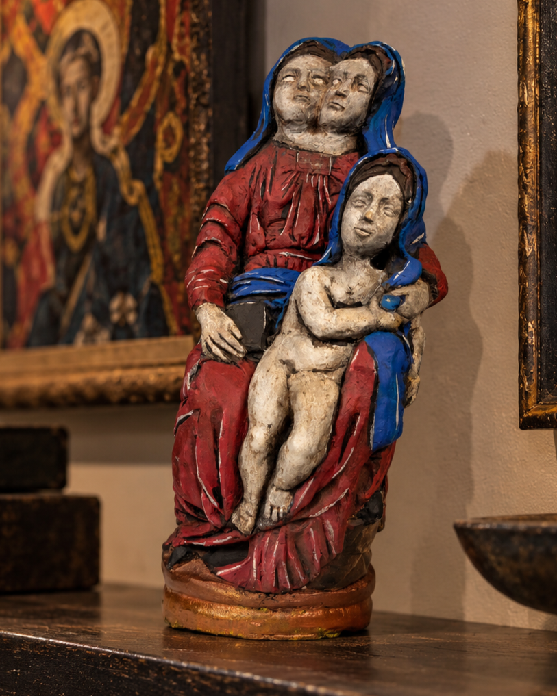 A painted sculpture of Madonna and Child, with Madonna holding a child against her chest, both with serene expressions. The sculpture is adorned with red and blue clothing and is displayed indoors with artwork in the background.