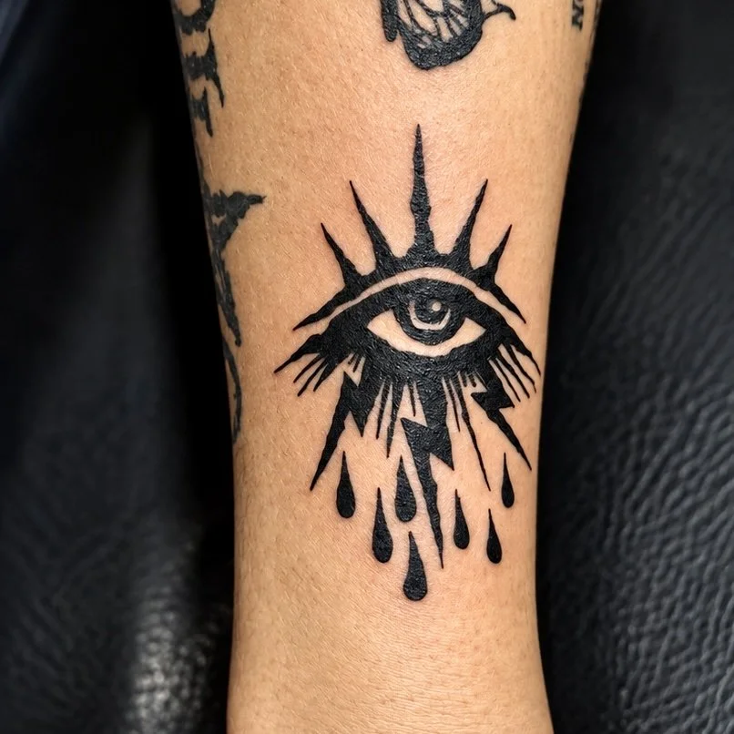 Tattoo of an eye with rays and teardrops, with lightning bolts beneath, in black ink.