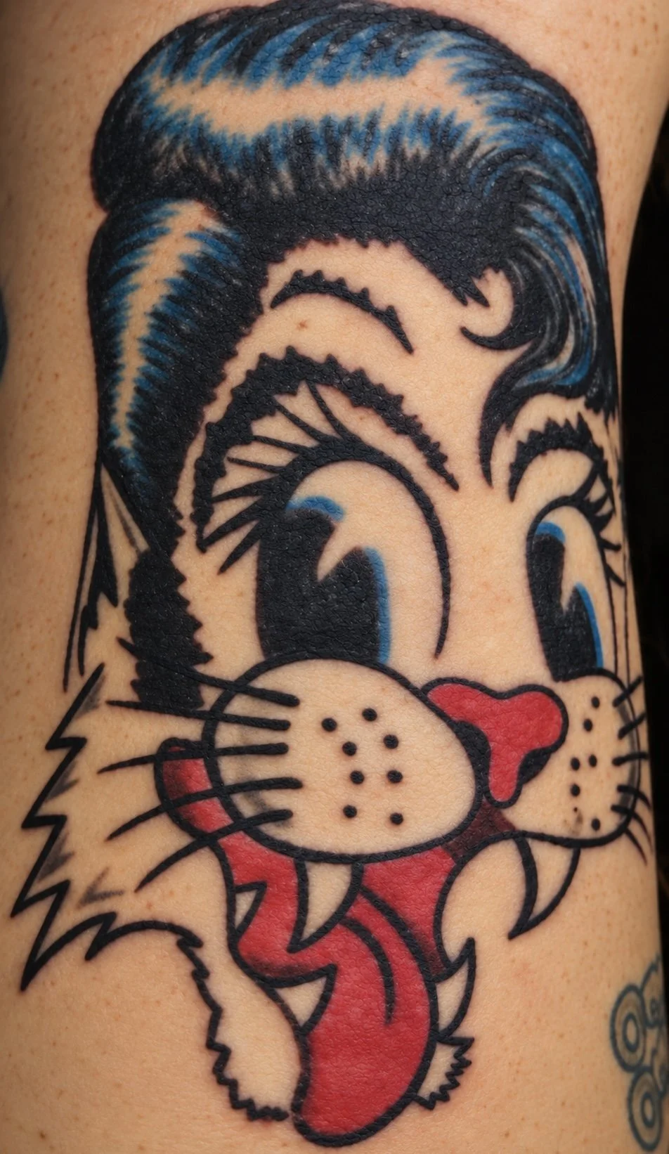 Tattoo of a cartoon cat with blue and black hair, big eyes, and a red tongue sticking out.