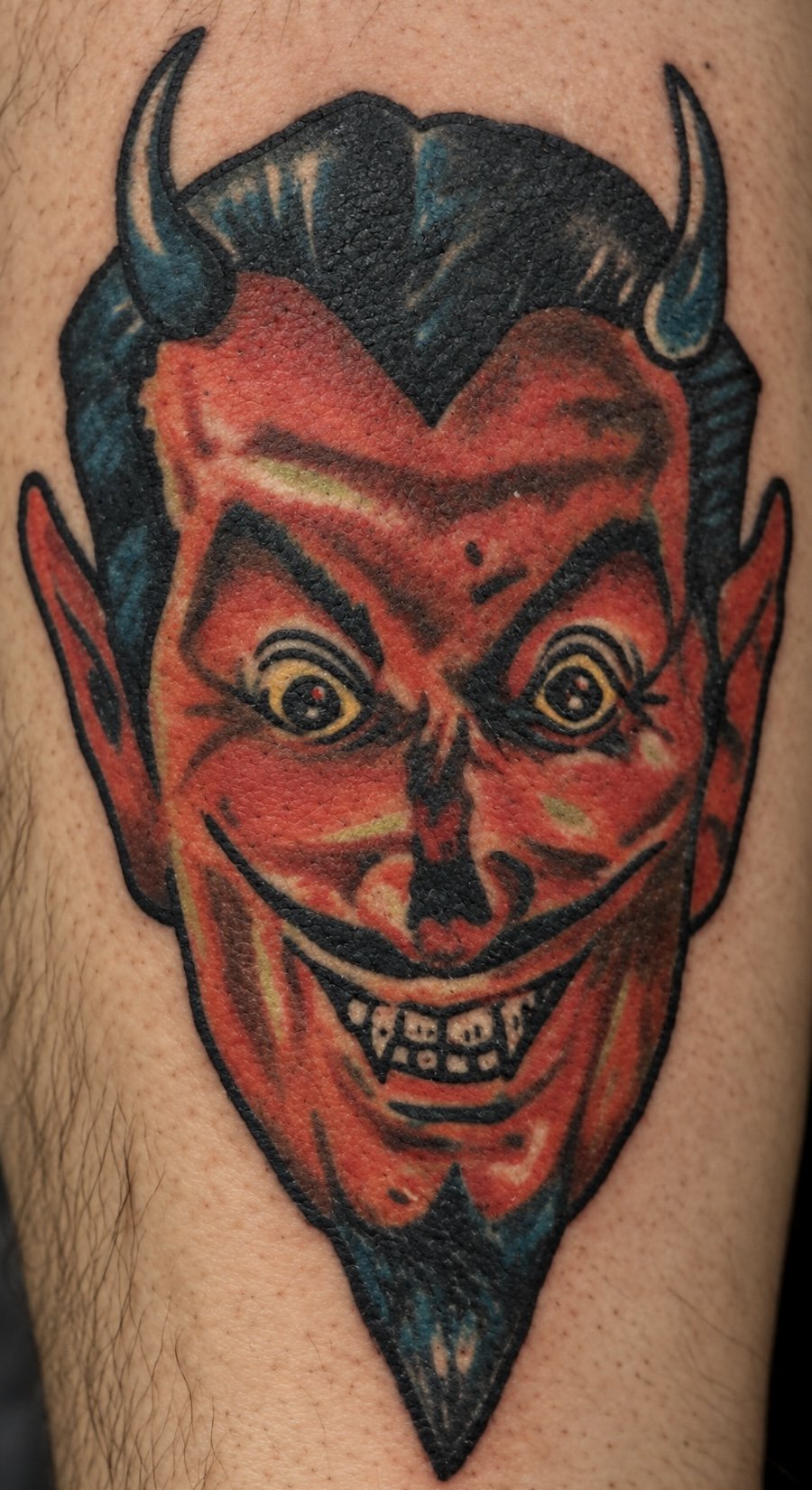 Tattoo of a smiling devil with red skin, pointed ears, black hair, yellow eyes, and blue horns.