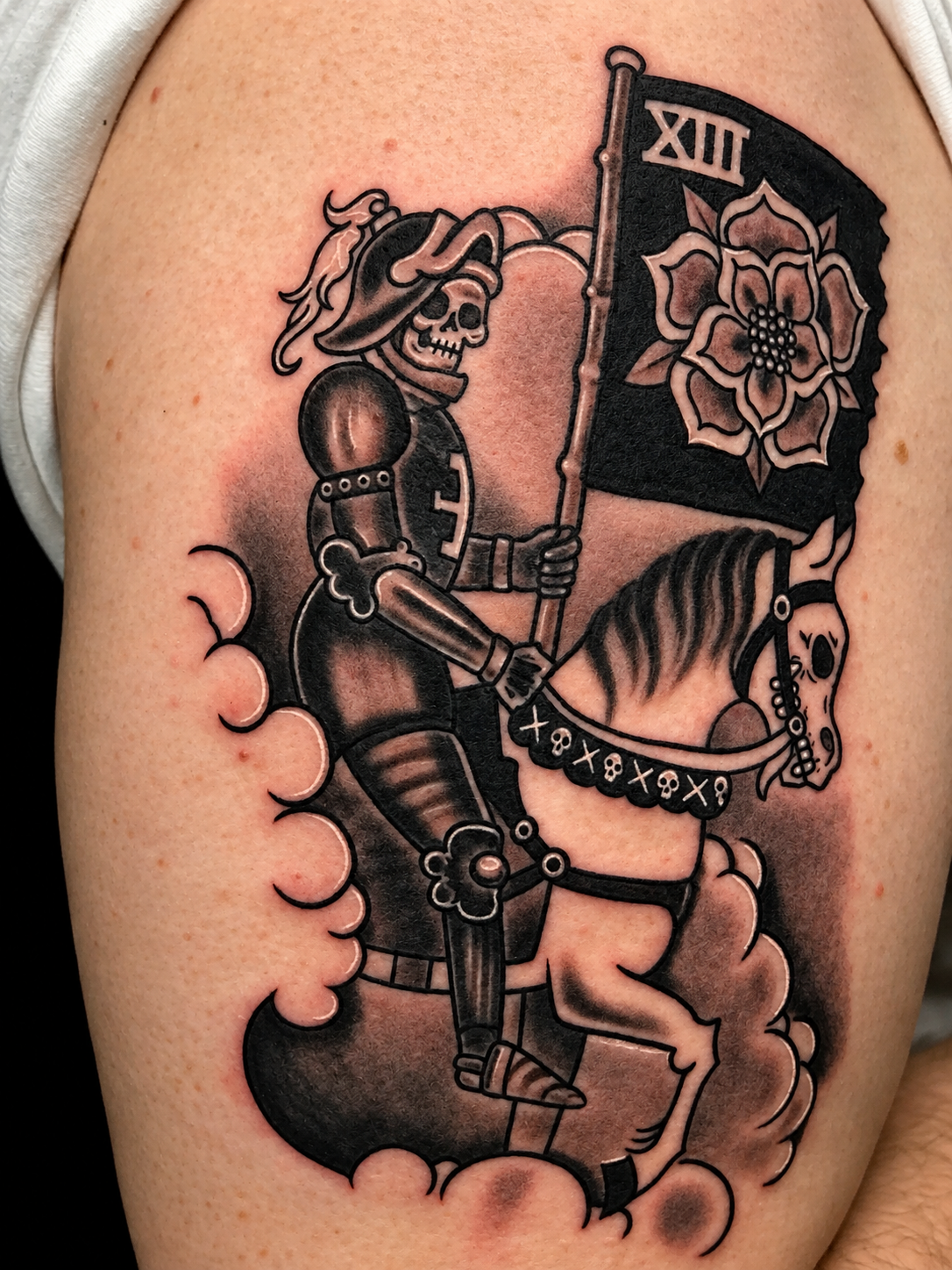 Tattoo of a skeleton knight riding a horse, holding a flag with a flower design, in black and gray ink.