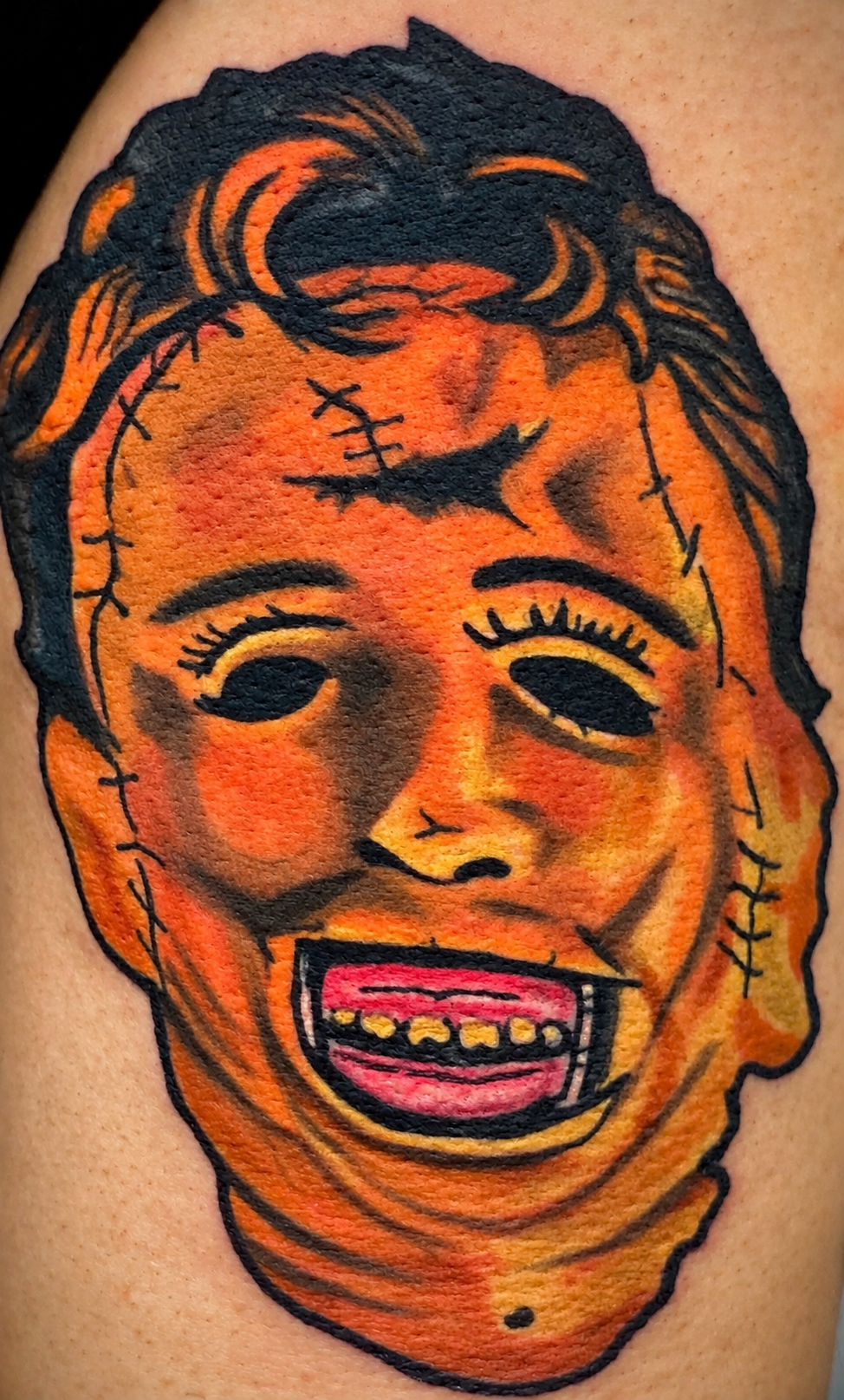 A colorful tattoo of a clown face with exaggerated features, including a wide open mouth showing gold teeth, dark eye sockets, and disheveled orange and black hair.