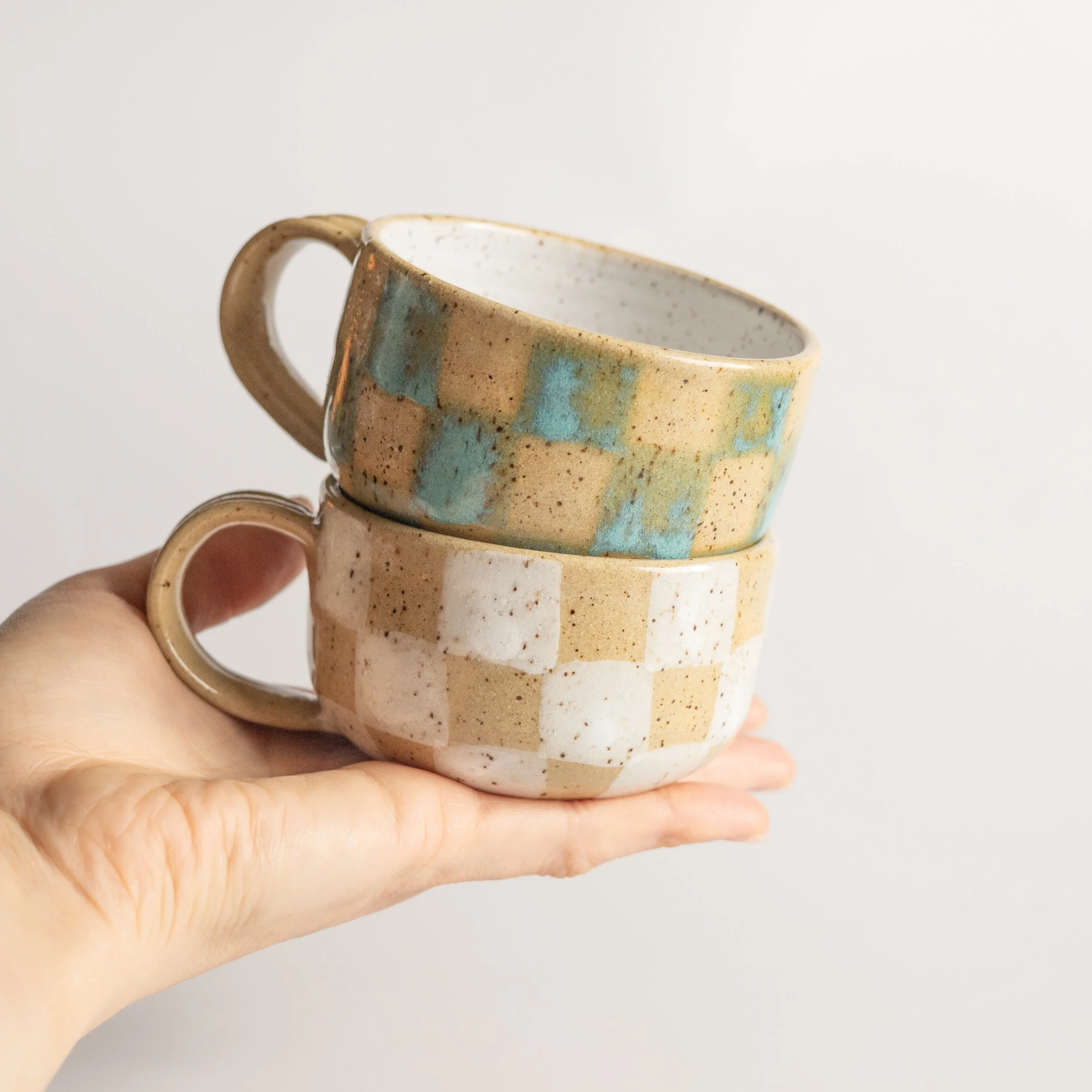 a hand holding two handmade ceramic mugs with a geometric checkered glaze design
