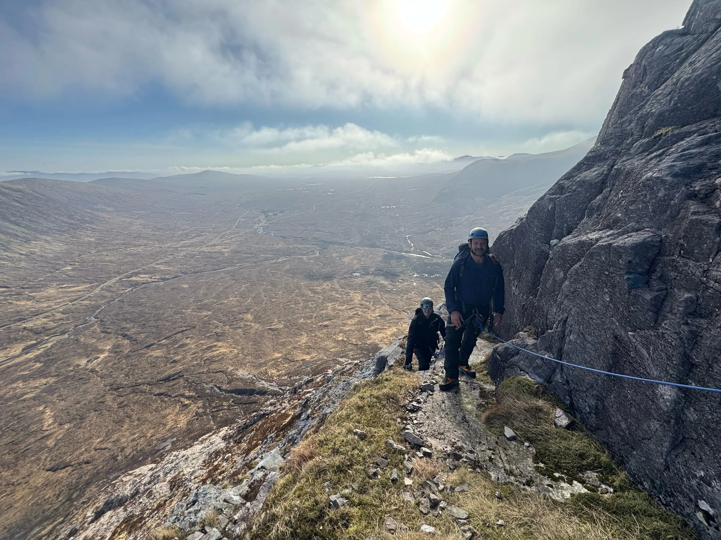 Two climbers with helmets and climbing gear ascending a steep mountain trail with a vast landscape in the background.
