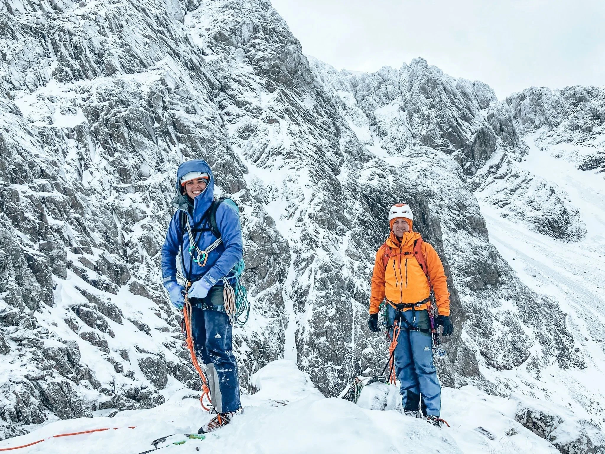 Two climbers dressed in colorful winter gear stand on snow-covered rocks at the base of a steep, snowy mountain with jagged rocks and cliffs.