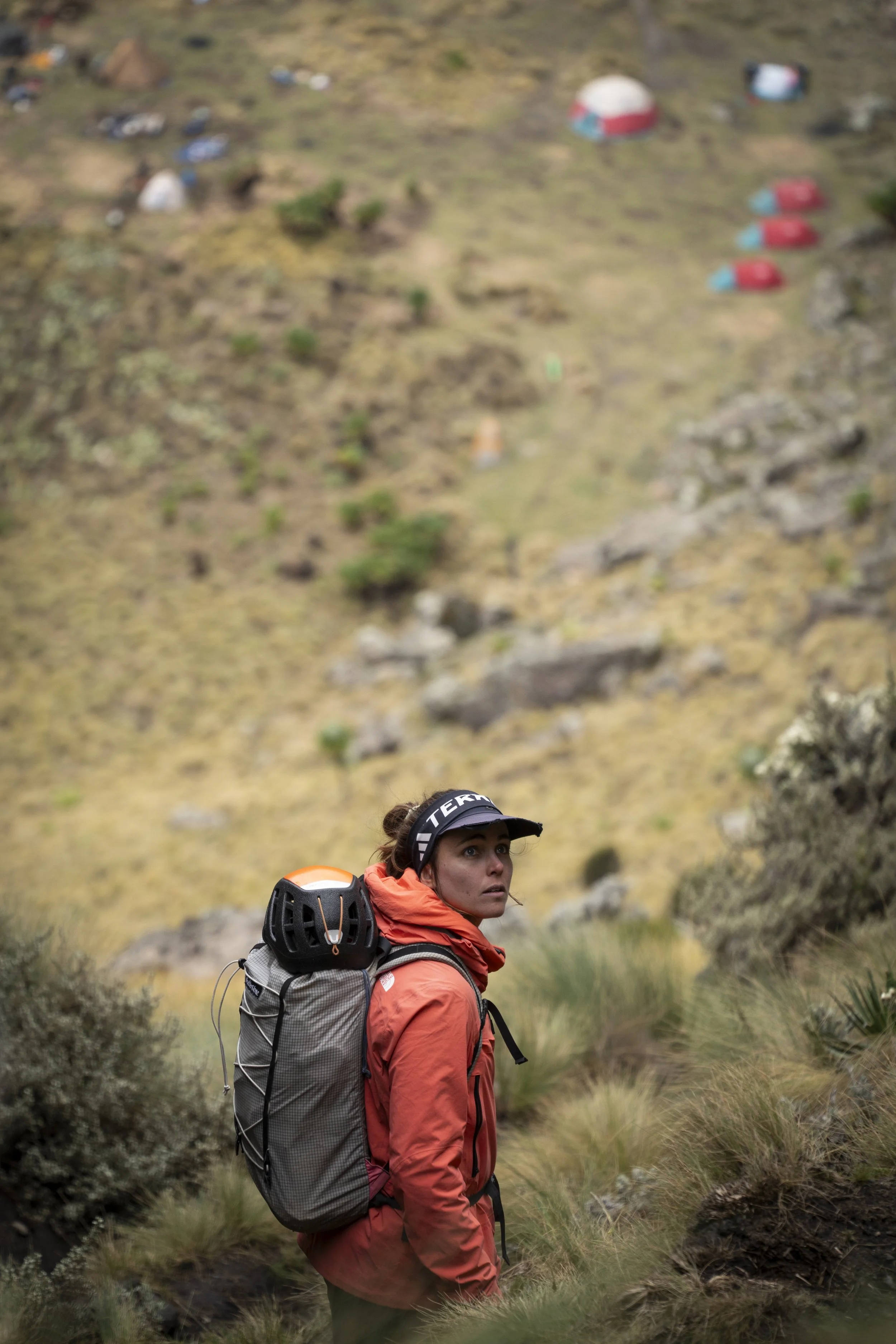A woman in an orange jacket with a backpack and helmet, standing outdoors on a grassy mountain trail, with a blurred background of tents and camping gear.