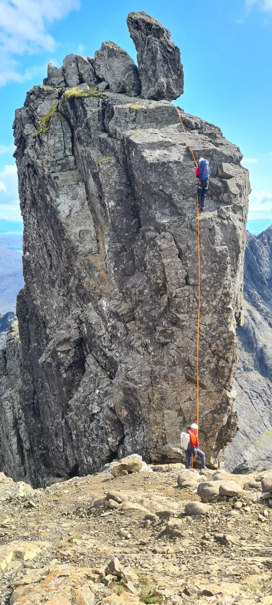 Two climbers ascending a large granite rock formation, with one near the bottom and the other higher up, connected by an orange rope, in a mountainous landscape under a partly cloudy sky.