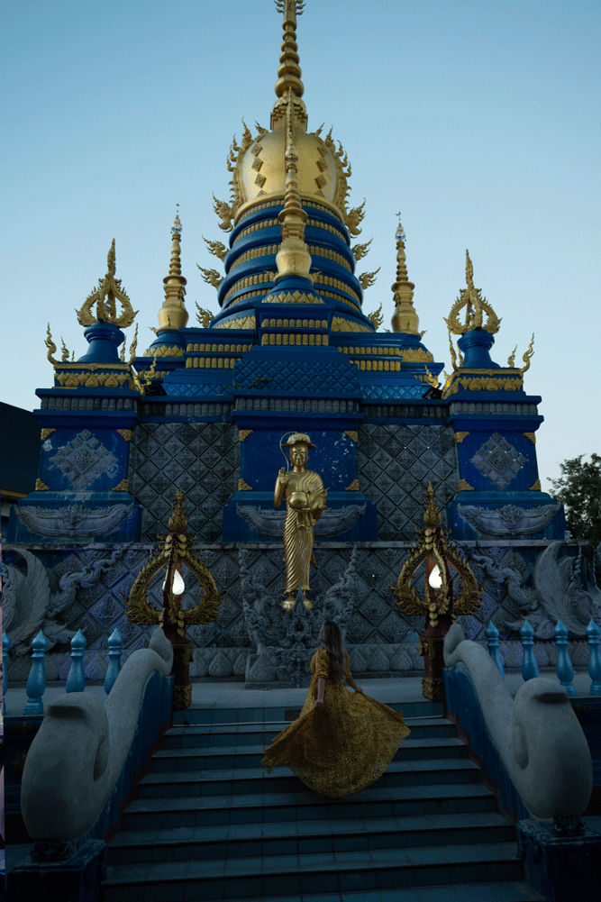 A woman in a gold dress standing on steps leading to an ornate blue and gold temple with statues and decorative elements.