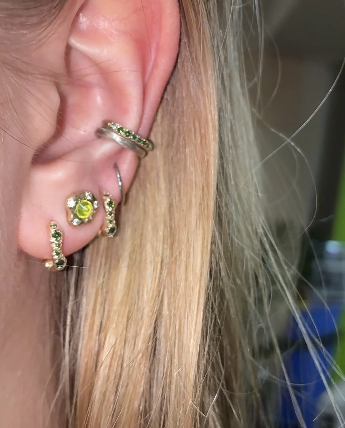 A woman's ear wearing various green hoops and studs, including 2 stacked ear cuffs
