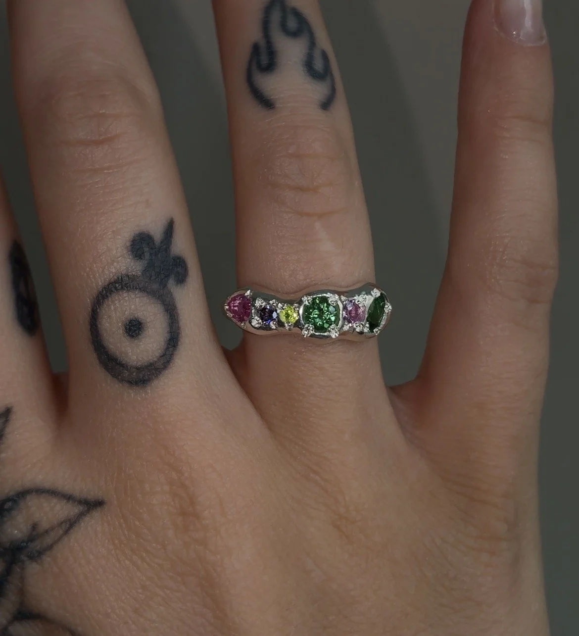 A molten silver ring with multicoloured gemstones photographed on a hand