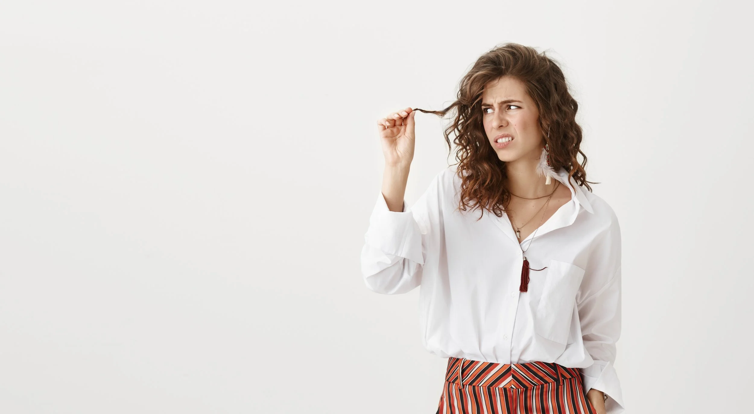 Mulher de cabelo castanho encaracolado observa uma madeixa de cabelo com expressão de preocupação, ilustrando dúvidas sobre queda ou quebra capilar.