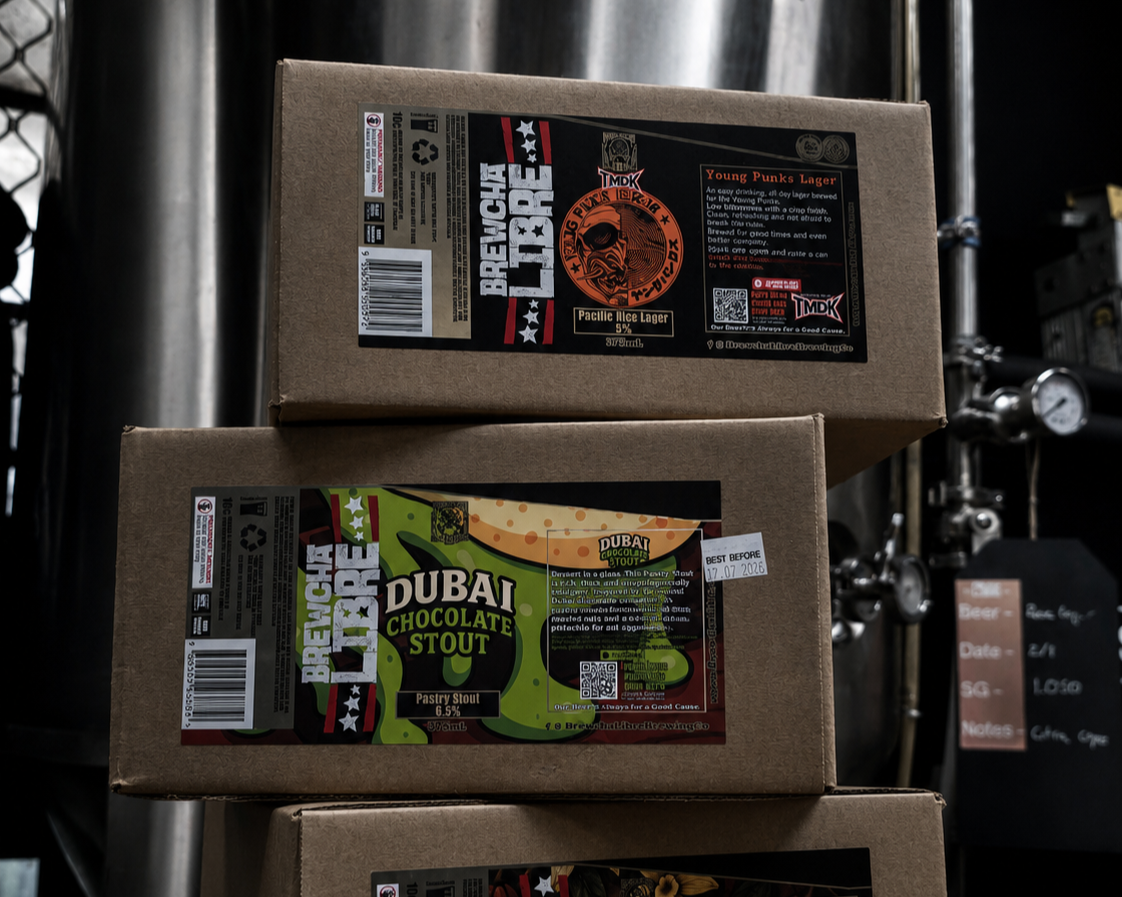 Three cardboard boxes of beer stacked on each other in a brewery, with labels indicating different flavors such as 'Breloca Hare', 'Dubai Chocolate Stout', and 'Young Punks Lager'.