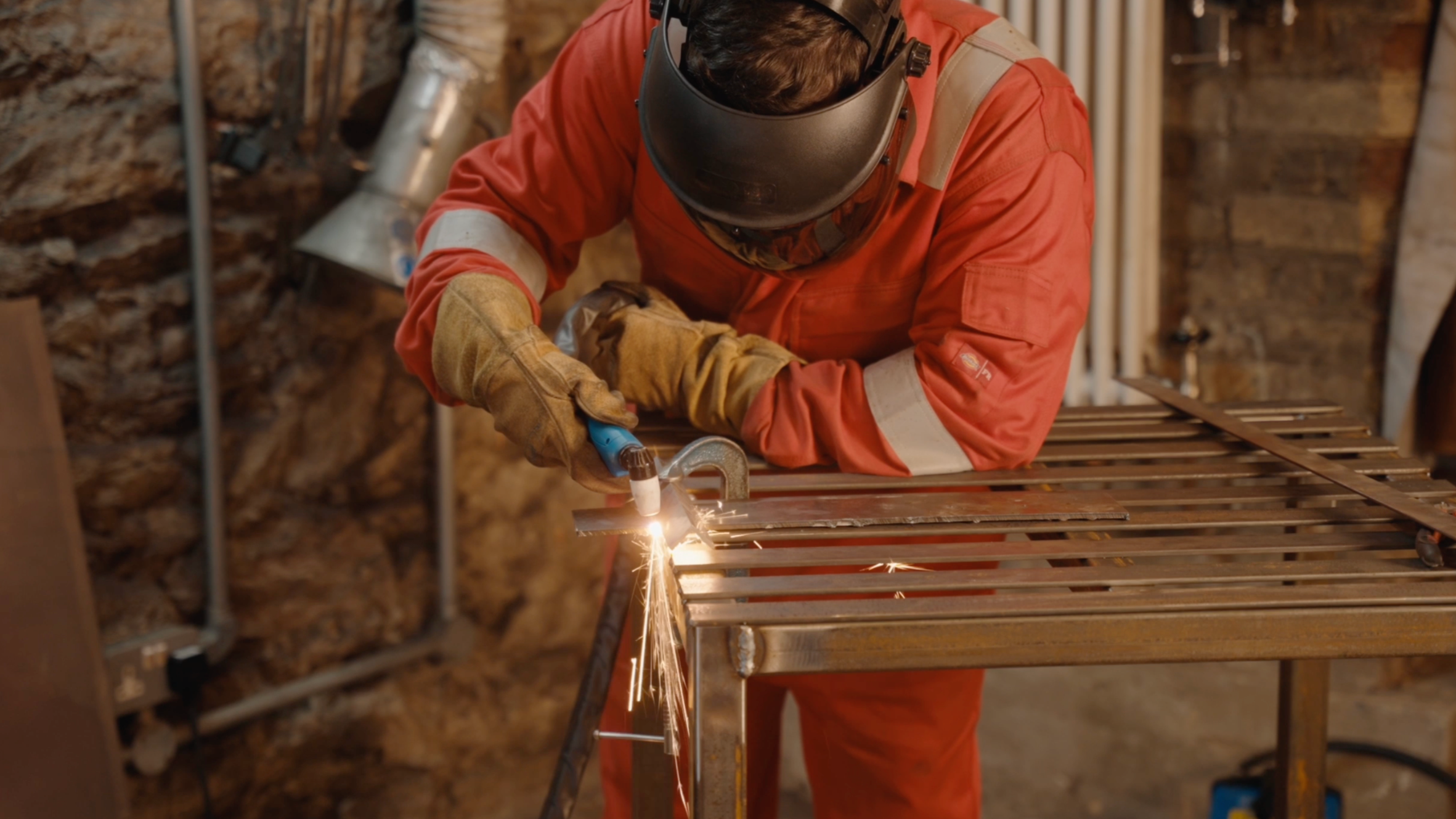 A person welding metal in a workshop, wearing protective gear including a welding helmet and gloves, and working on a metal frame with sparks flying.