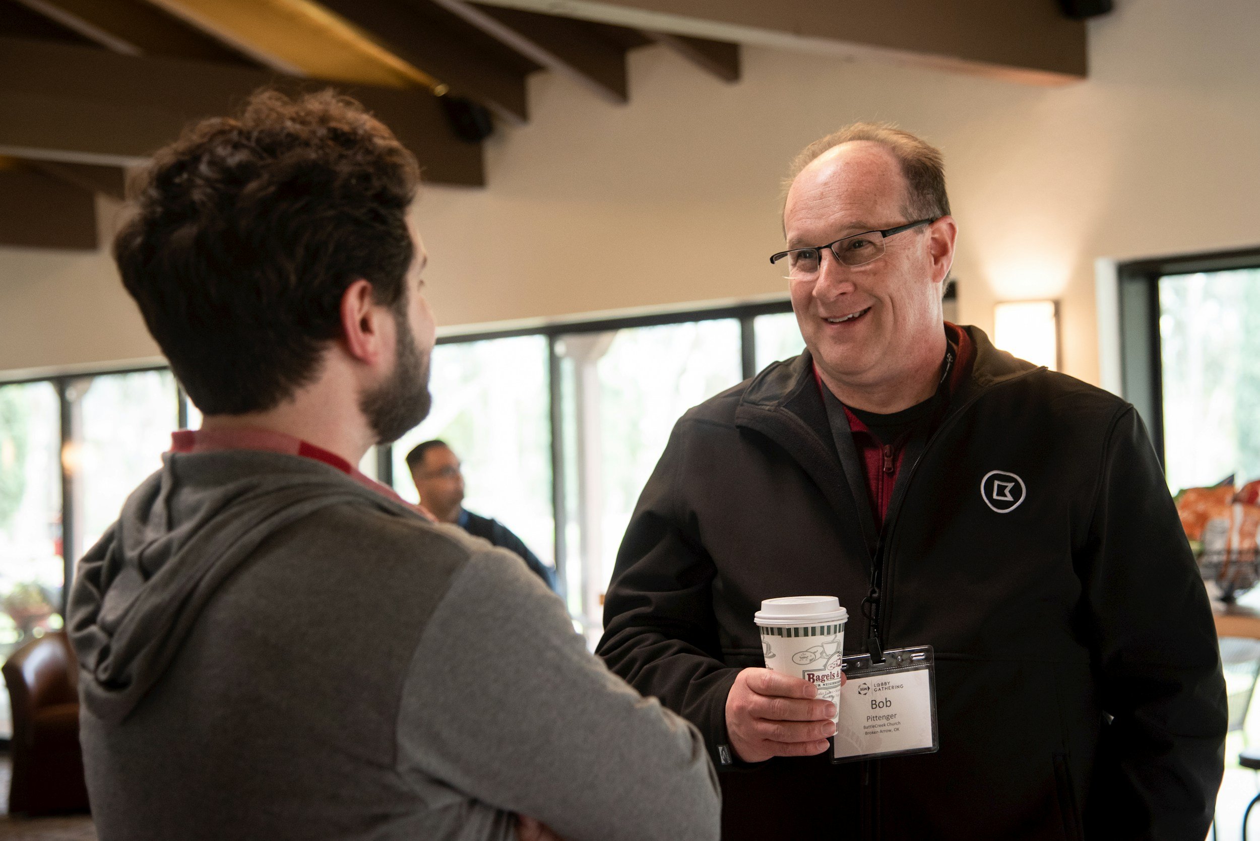 Two men are talking and smiling during a coffee break at a conference. One man is holding a coffee cup and wearing a conference badge that says Bob. The setting appears to be a cafe or lounge area with large windows and natural light.
