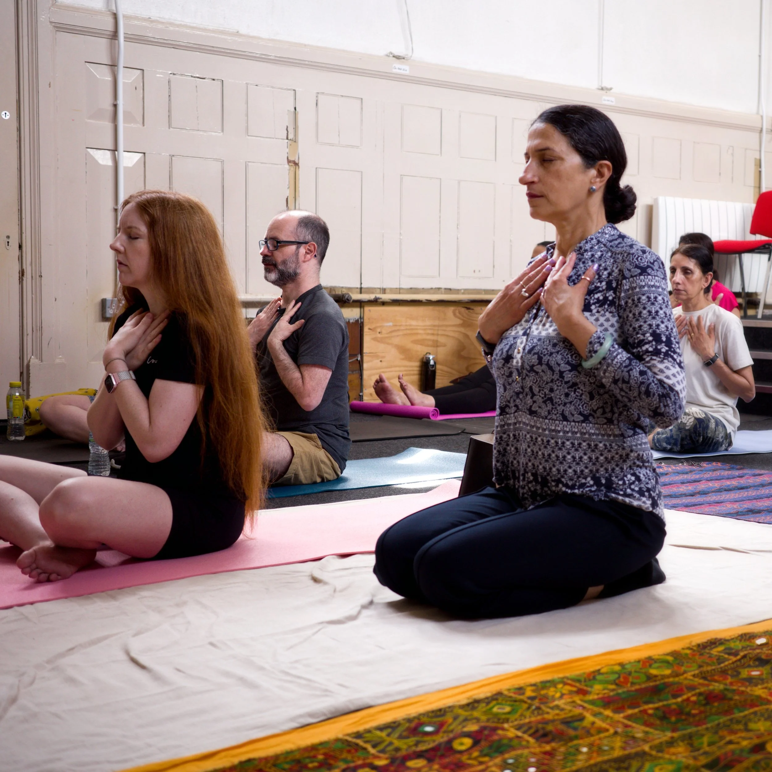 Group of people practicing yoga in a seated position, with hands on their chests, on yoga mats indoors.
