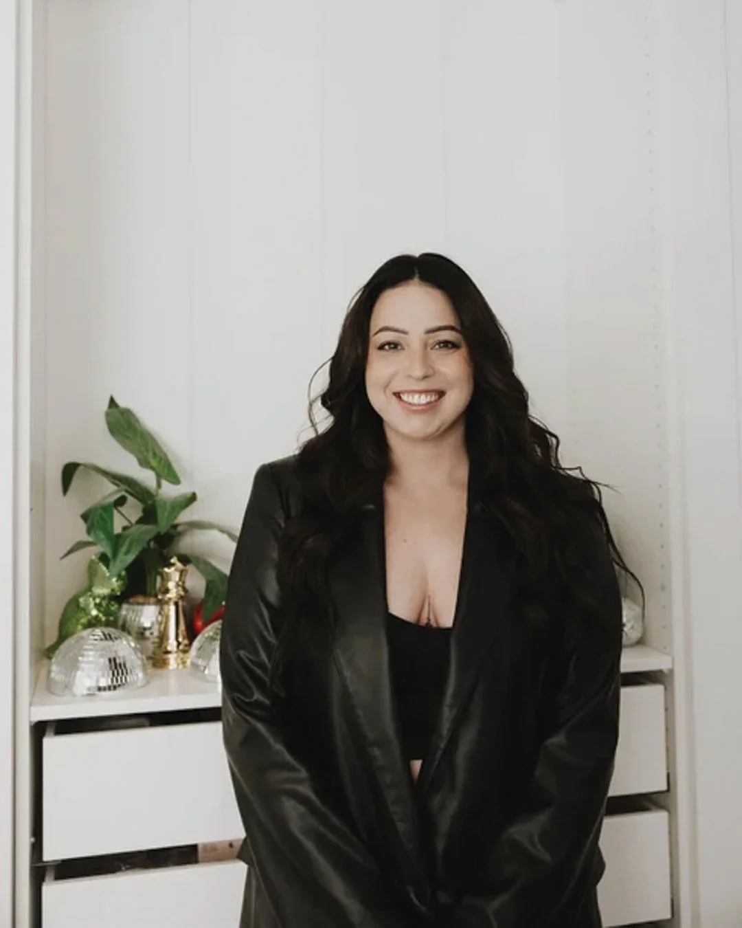 A woman with long dark wavy hair smiling, wearing a black leather jacket and black top, standing in front of a white wall with a white dresser, a green plant, and decorative household items in the background.
