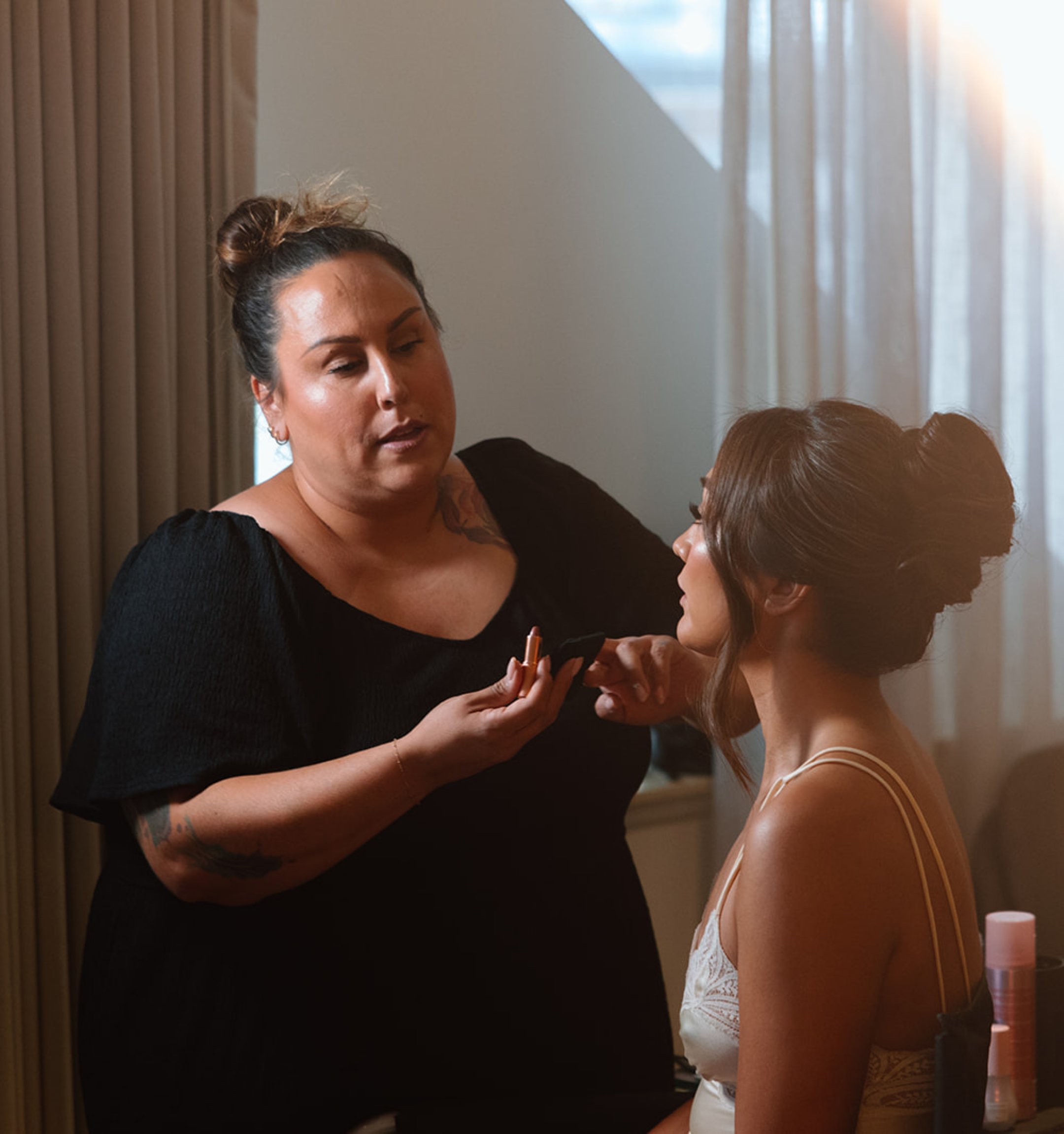 A makeup artist is applying makeup to a woman with styled hair in a softly lit room.