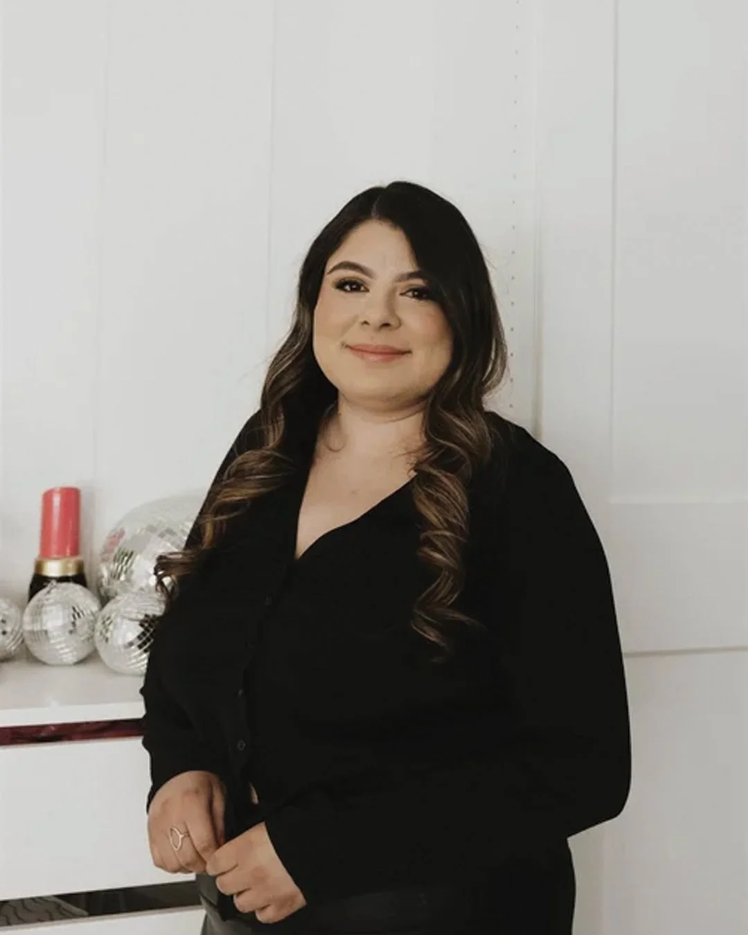 A woman with long wavy dark hair and light skin, smiling, wearing a black shirt, standing indoors with white walls and decorations including metallic disco balls and a red and gold bottle or candle in the background.