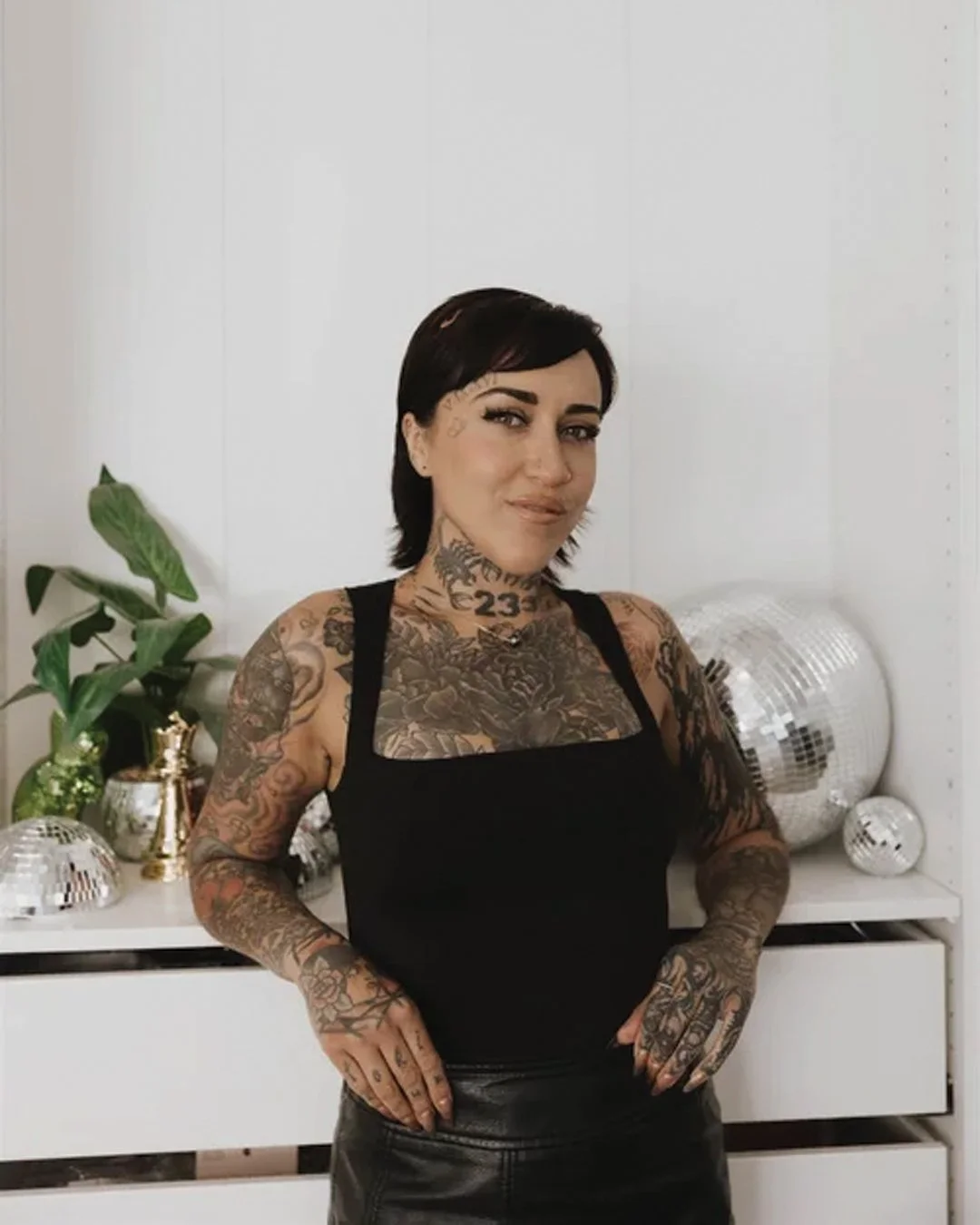 A woman with short dark hair and numerous tattoos on her arms and neck, wearing a black sleeveless top, standing in front of a white wall with decorative disco balls and a plant.