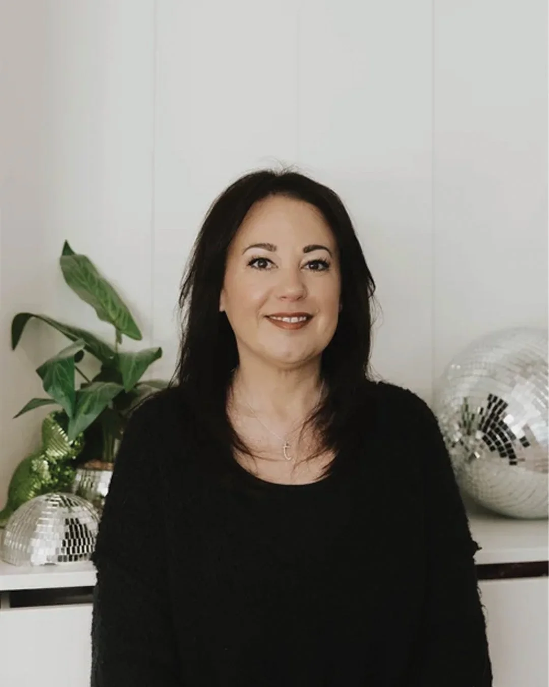 A woman with dark hair wearing a black top, smiling, standing in front of a white wall with two disco balls and a green plant.