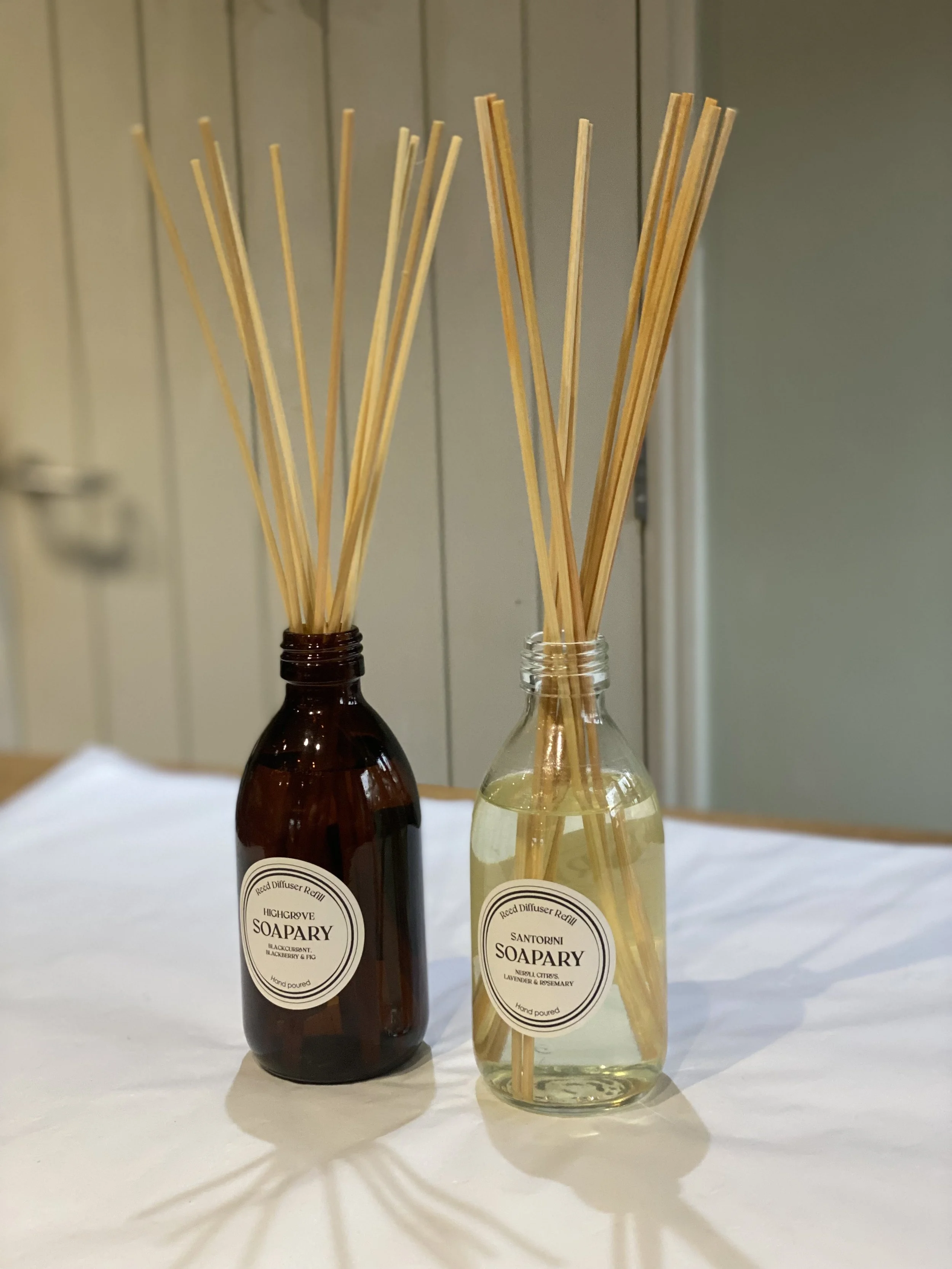 Two small bottles with reed diffusers, one dark brown labeled 'Highgrose Soapary' and the other clear labeled 'Santorini Soapary', with wooden sticks inserted into each bottle, on a white surface.