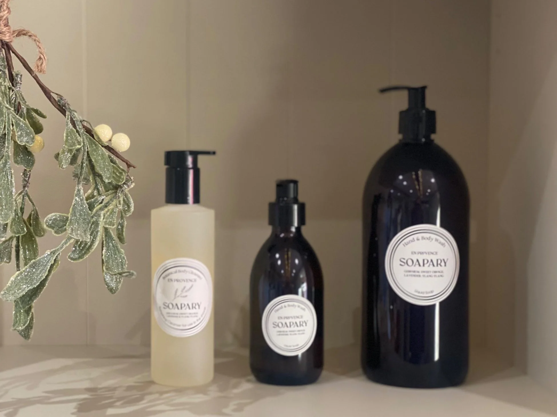 Three soap dispensers on a shelf with a frosted branch with leaves and berries on the left side.