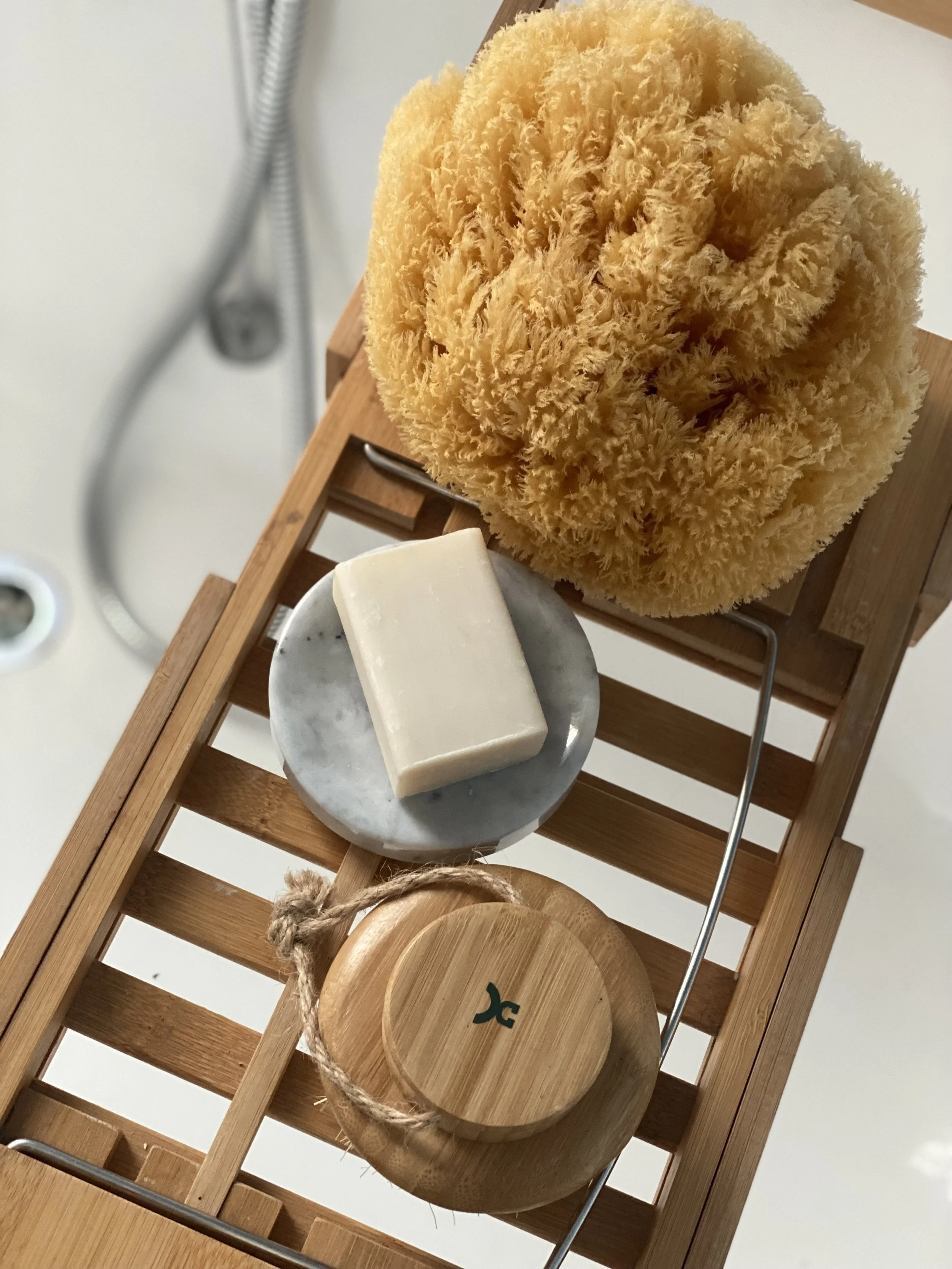 A beige natural sea sponge, a bar of soap on a marble soap dish, and a round wooden container with a lid on a wooden slatted shelf.
