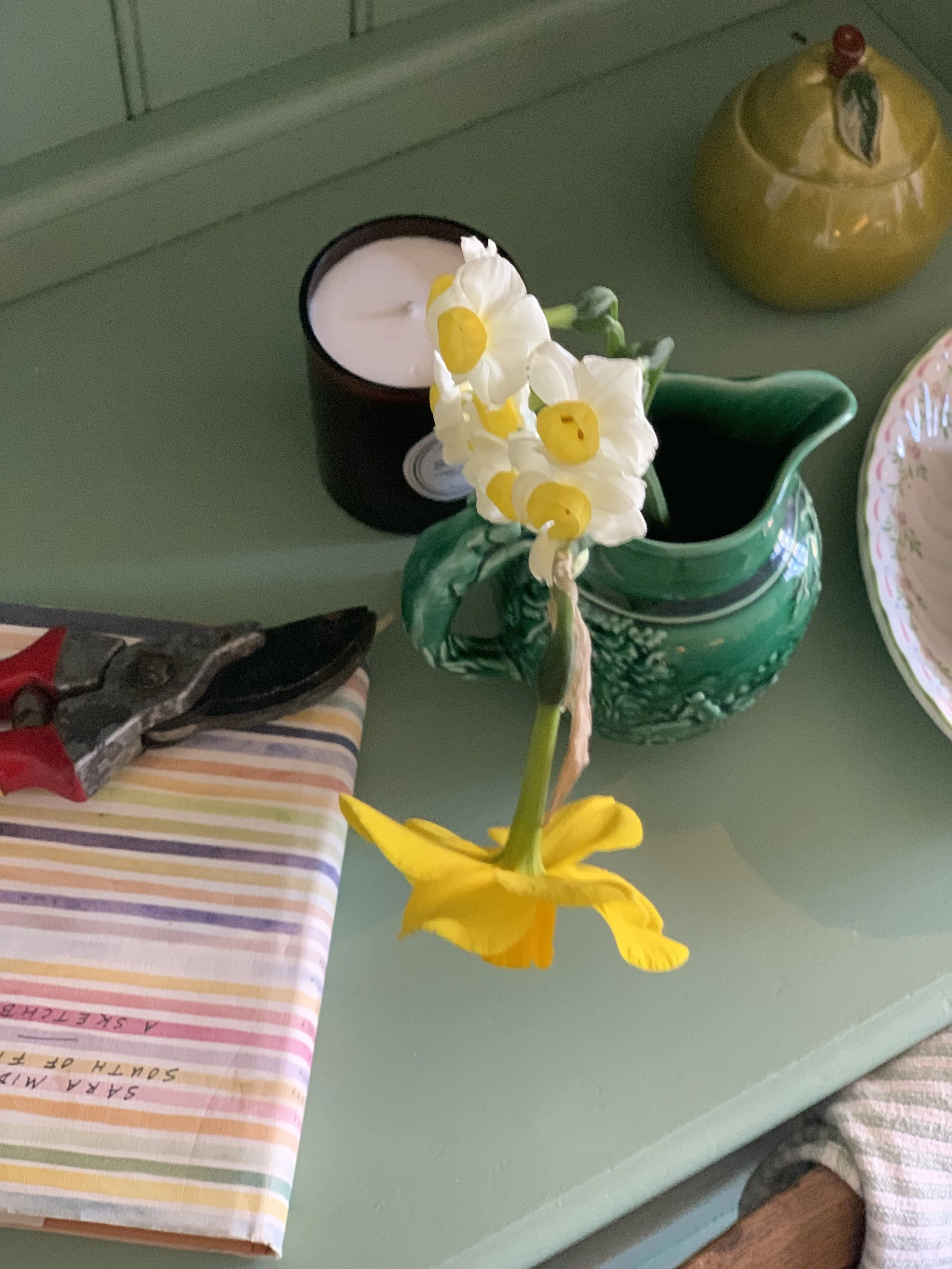 A decorative green vase with white and yellow flowers, a candle in a black holder, a yellow ceramic jar, and a patterned plate on a green table.